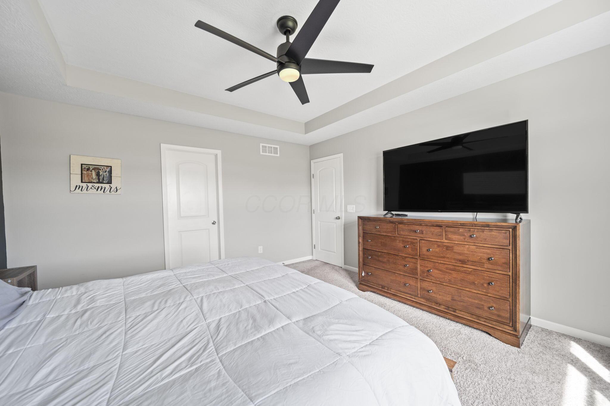 Bedroom with a white bed, tray ceiling, black fan, and TV on a wooden dresser.