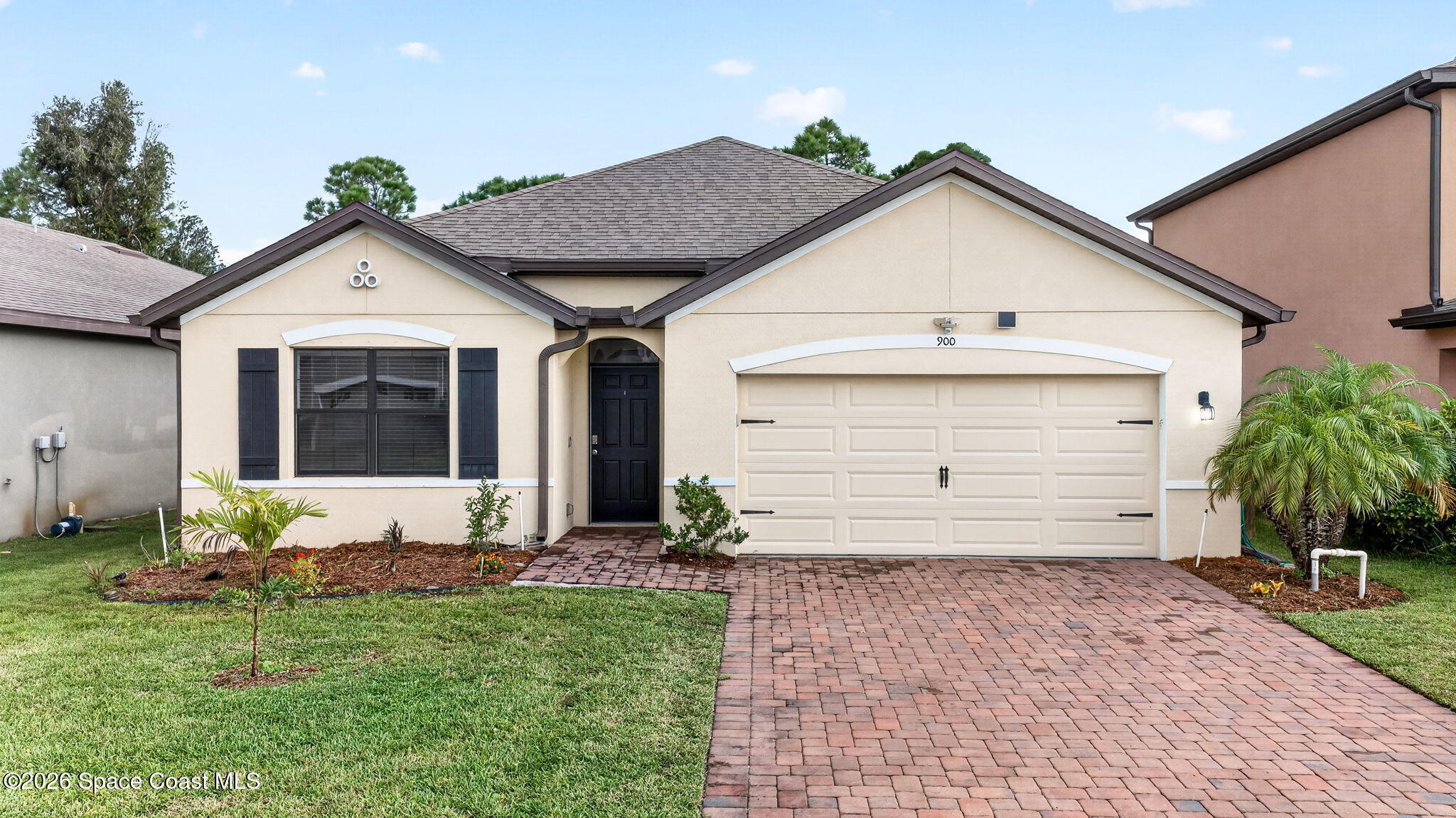 Beige single-story home with a two-car garage, brick paver driveway, and tropical landscaping.