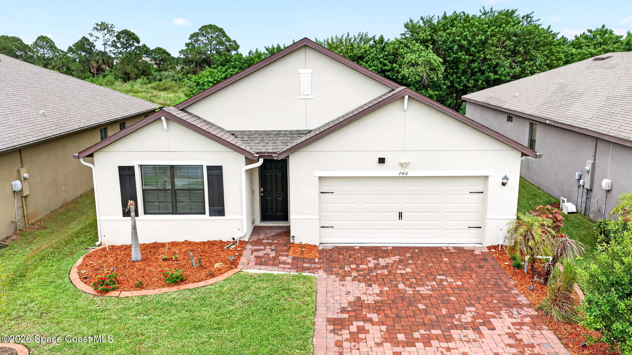 White single-story home with a two-car garage, brick paver driveway, and landscaped front beds.