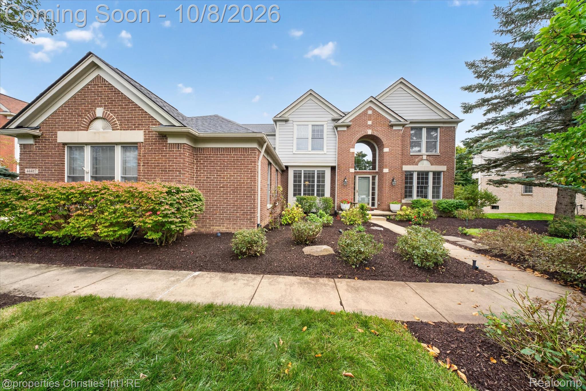 Brick-and-siding home with an arched entry, landscaped shrubs, and a curved walkway.