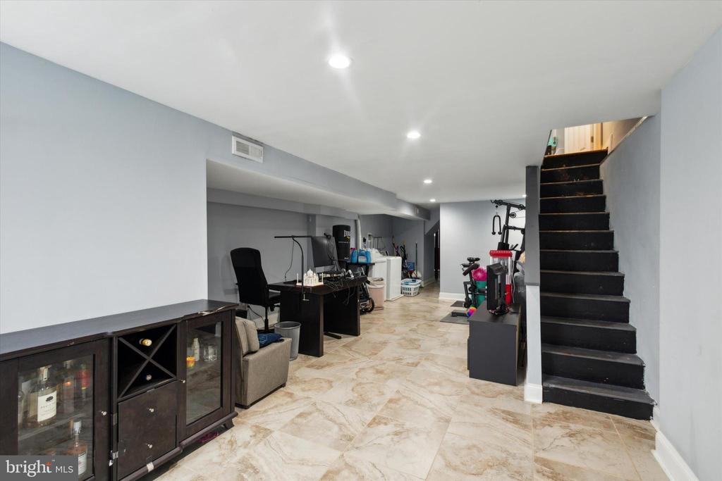 Finished basement with tiled floors, a dark staircase, exercise equipment and a desk.