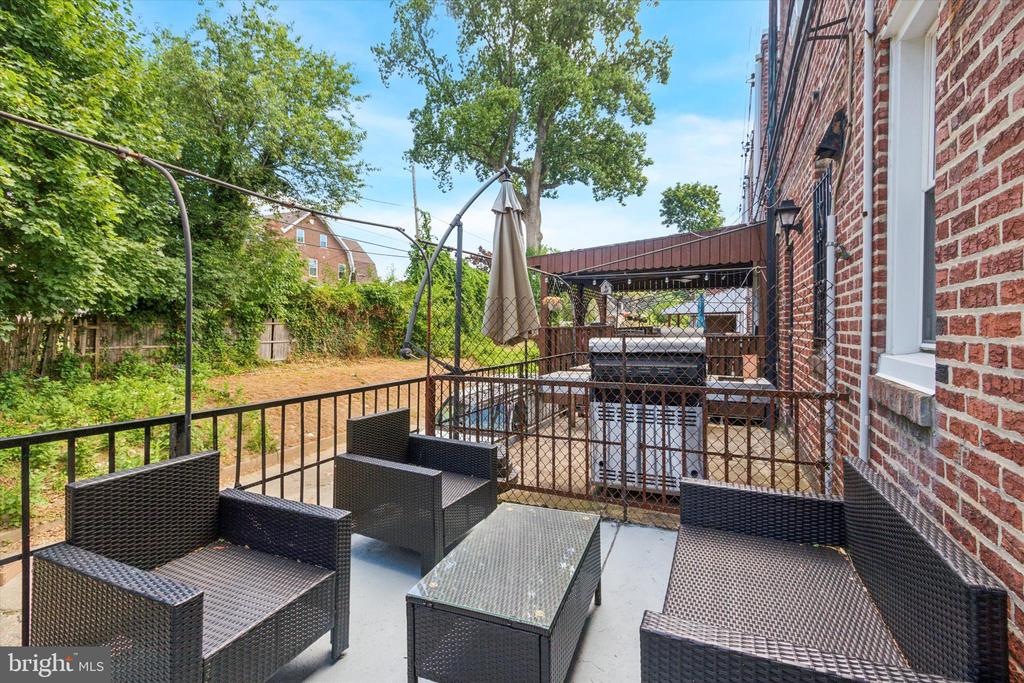 Furnished patio beside a brick house, with wicker seating and a covered deck overlooking a leafy yard.