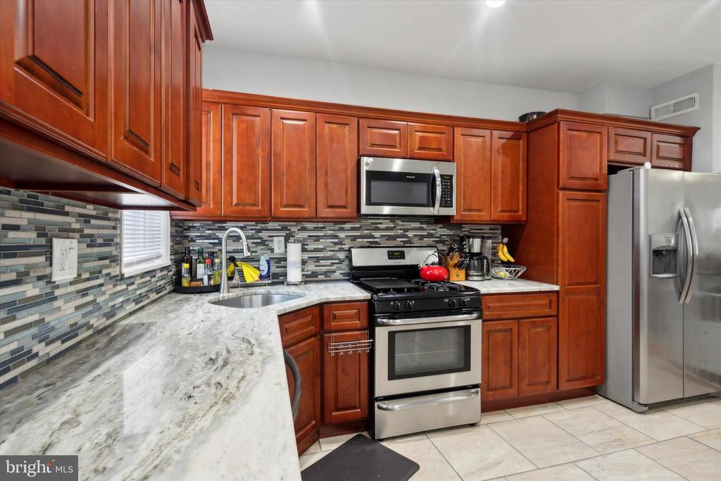 Kitchen with reddish-brown cabinetry, mosaic tile backsplash, stone counters, and stainless steel appliances.