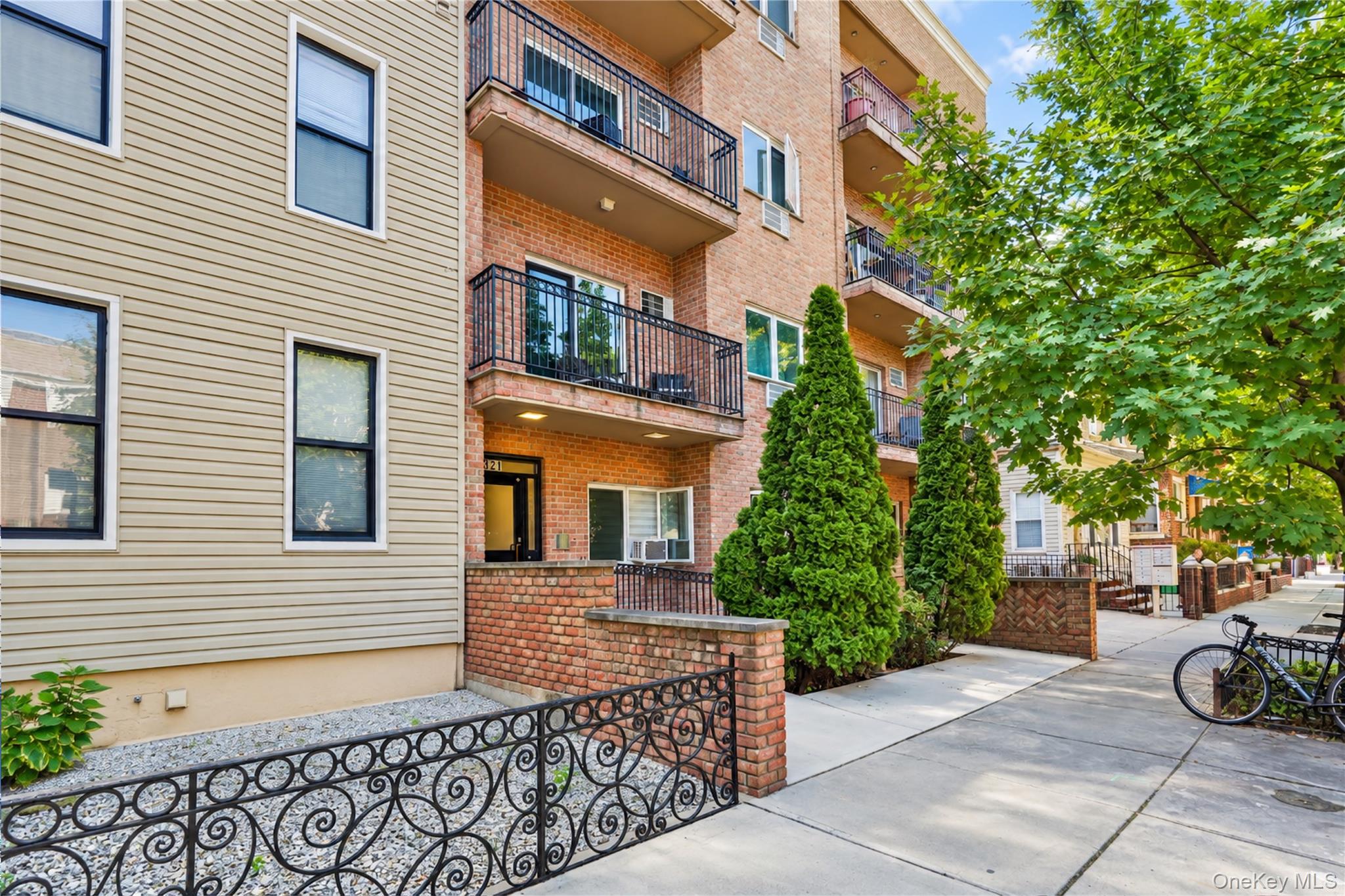 Apartment building with brick facade, black-railed balconies, and landscaped entrance along a tree-lined sidewalk.