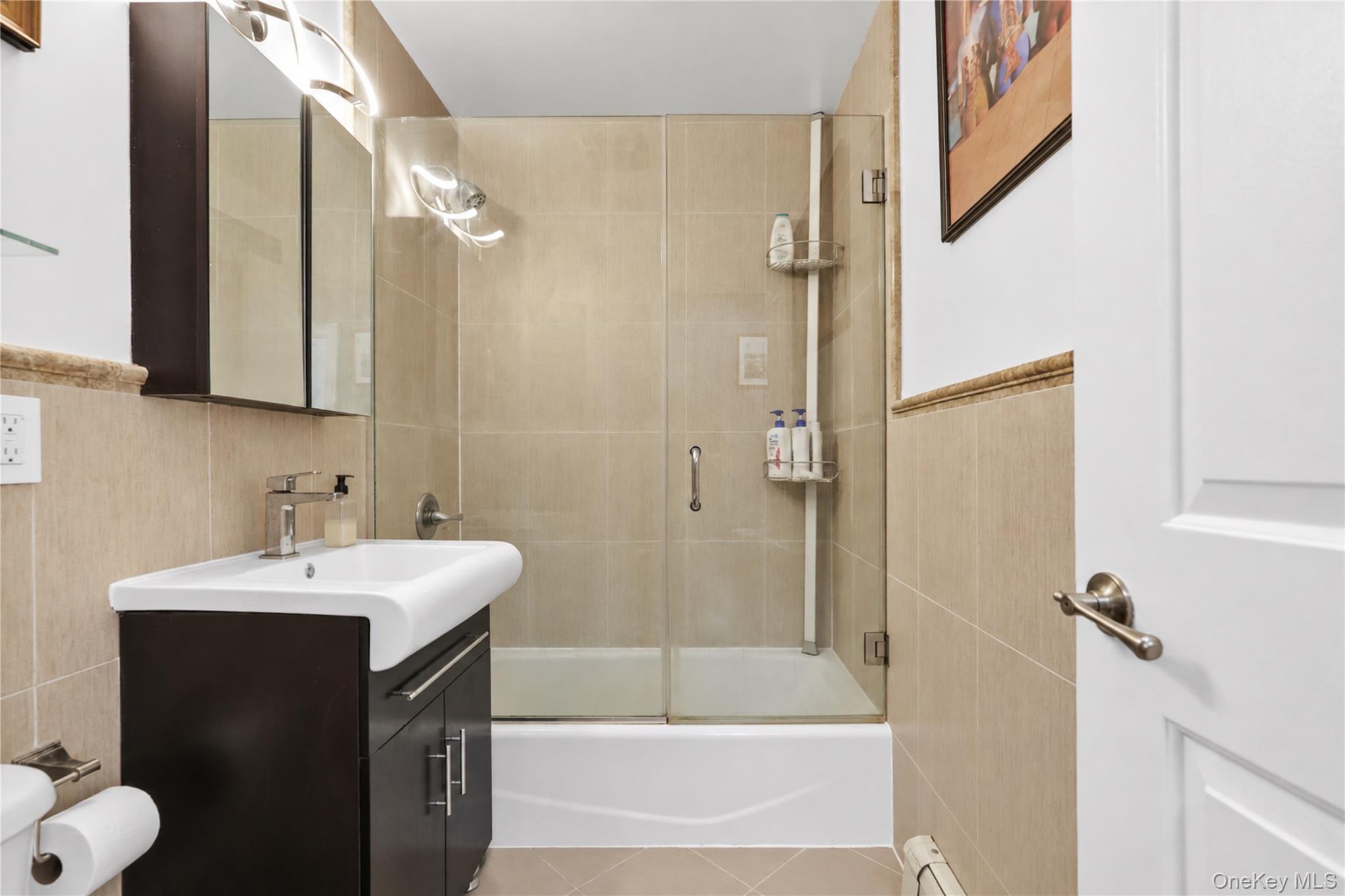 Bathroom with a glass-enclosed tub and beige tile beside a dark vanity.