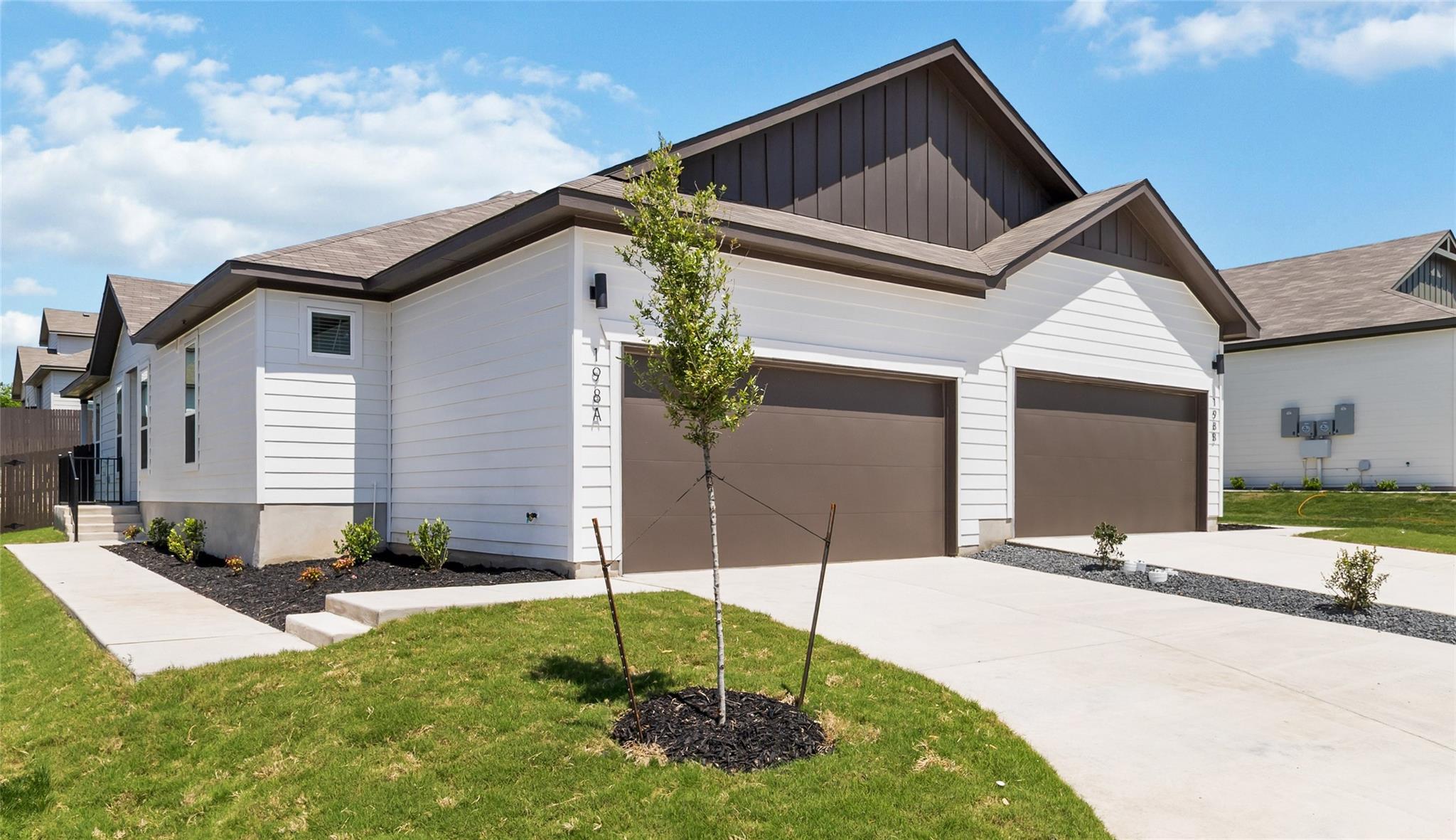 White home with two brown garage doors, dark gabled roofs, and a young tree in the front yard.