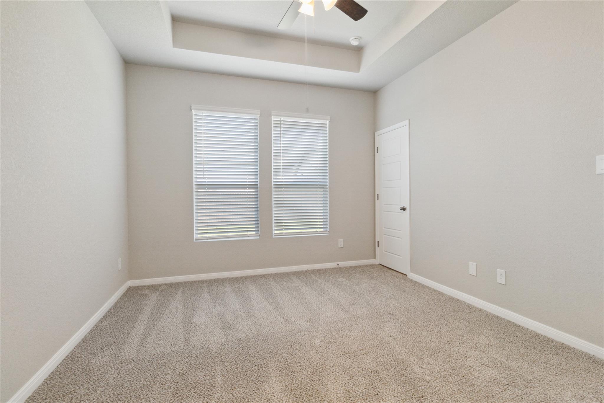 Empty bedroom with beige carpet, two blind-covered windows, and a tray ceiling with ceiling fan.
