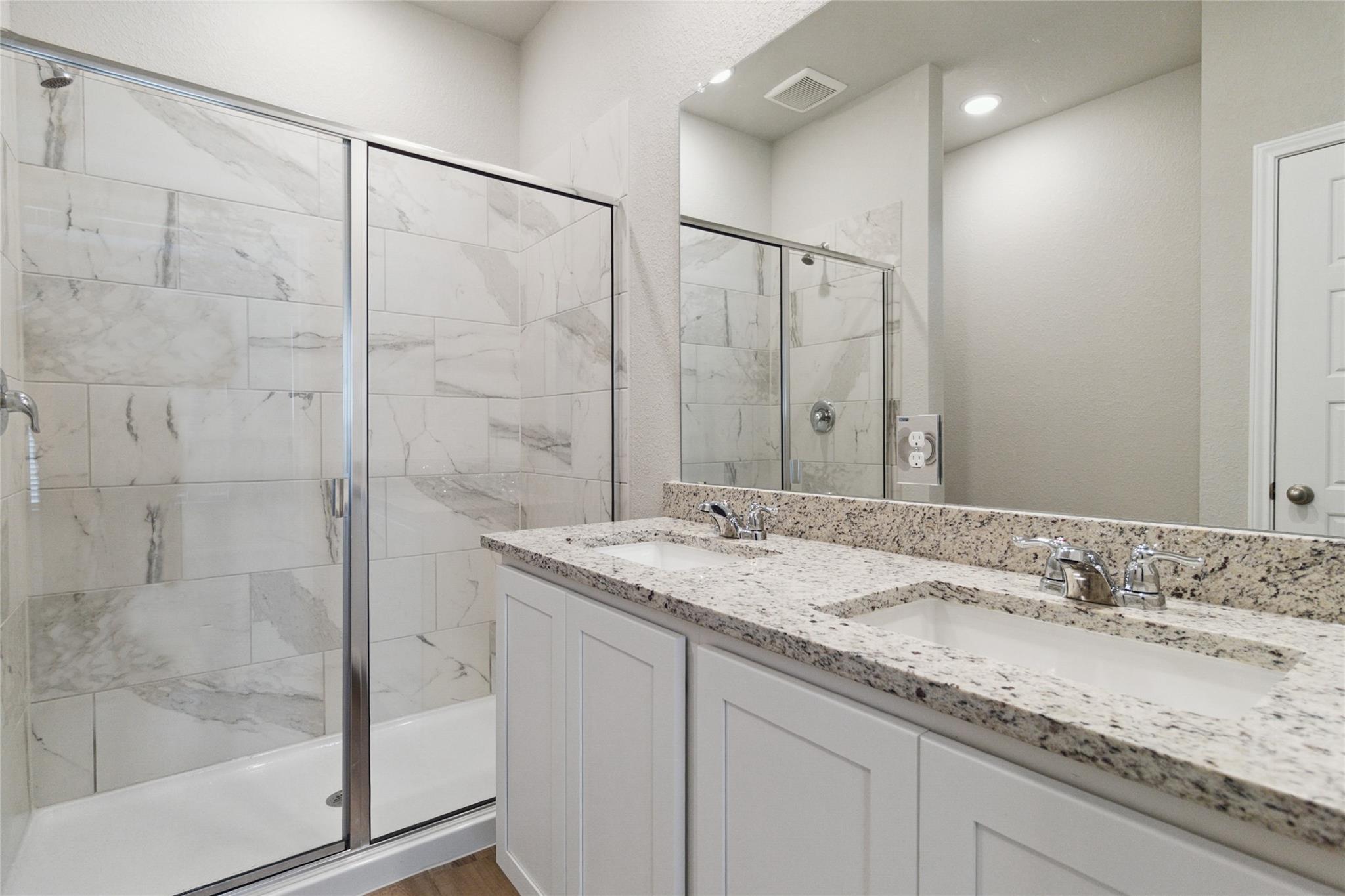 Bathroom with a marble-look tiled shower and double vanity topped with speckled granite.