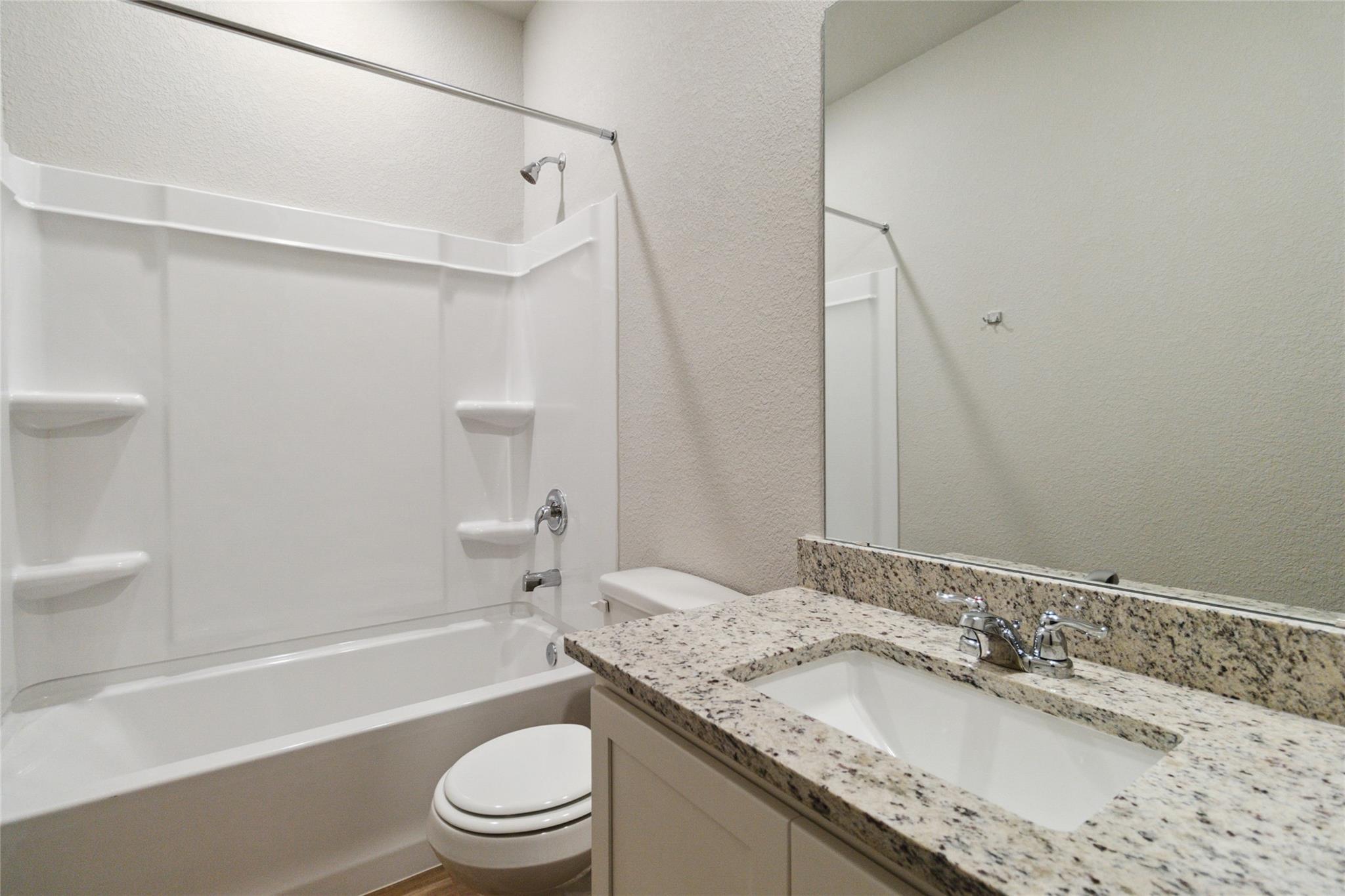 Bathroom with a granite vanity, inset sink, white tub and shower, and toilet.