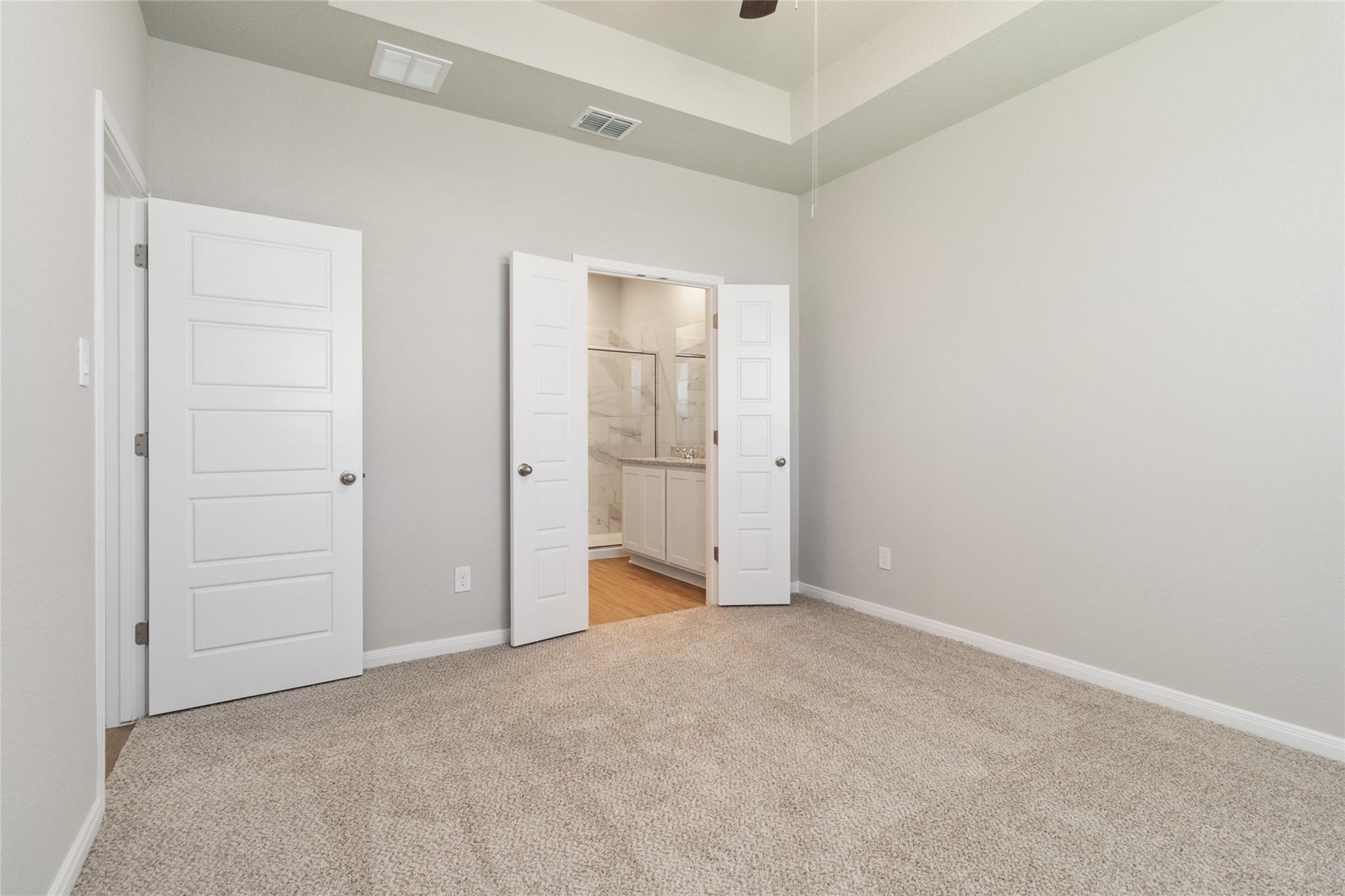 Empty carpeted bedroom with a tray ceiling and open doors to an ensuite bathroom.