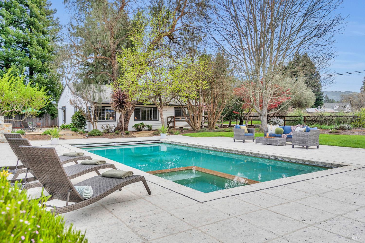 Backyard swimming pool beside a white house, with a stone patio and outdoor lounge seating.