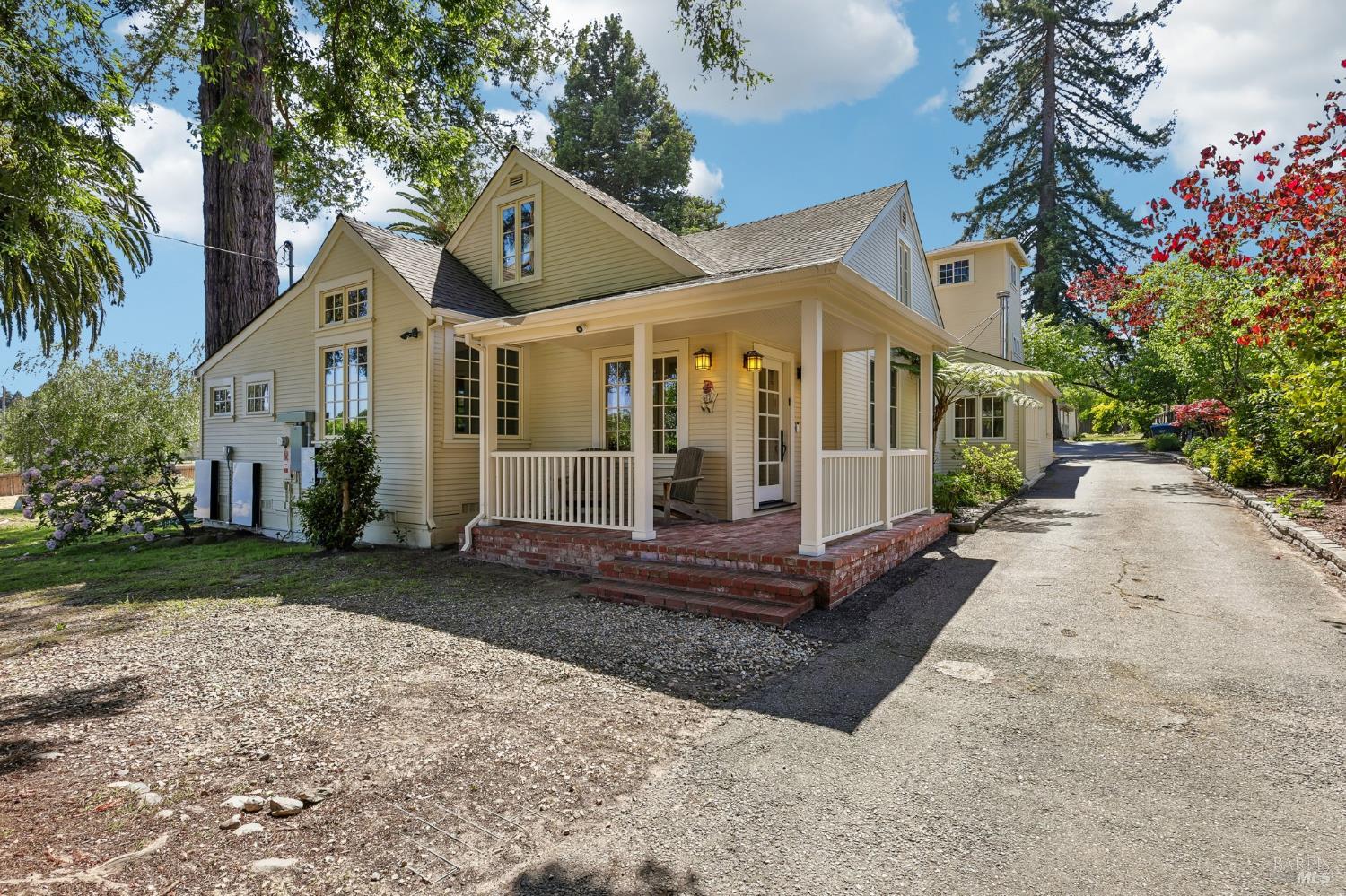 Cream-colored house with a covered porch, brick steps, and a gravel driveway beneath mature trees.