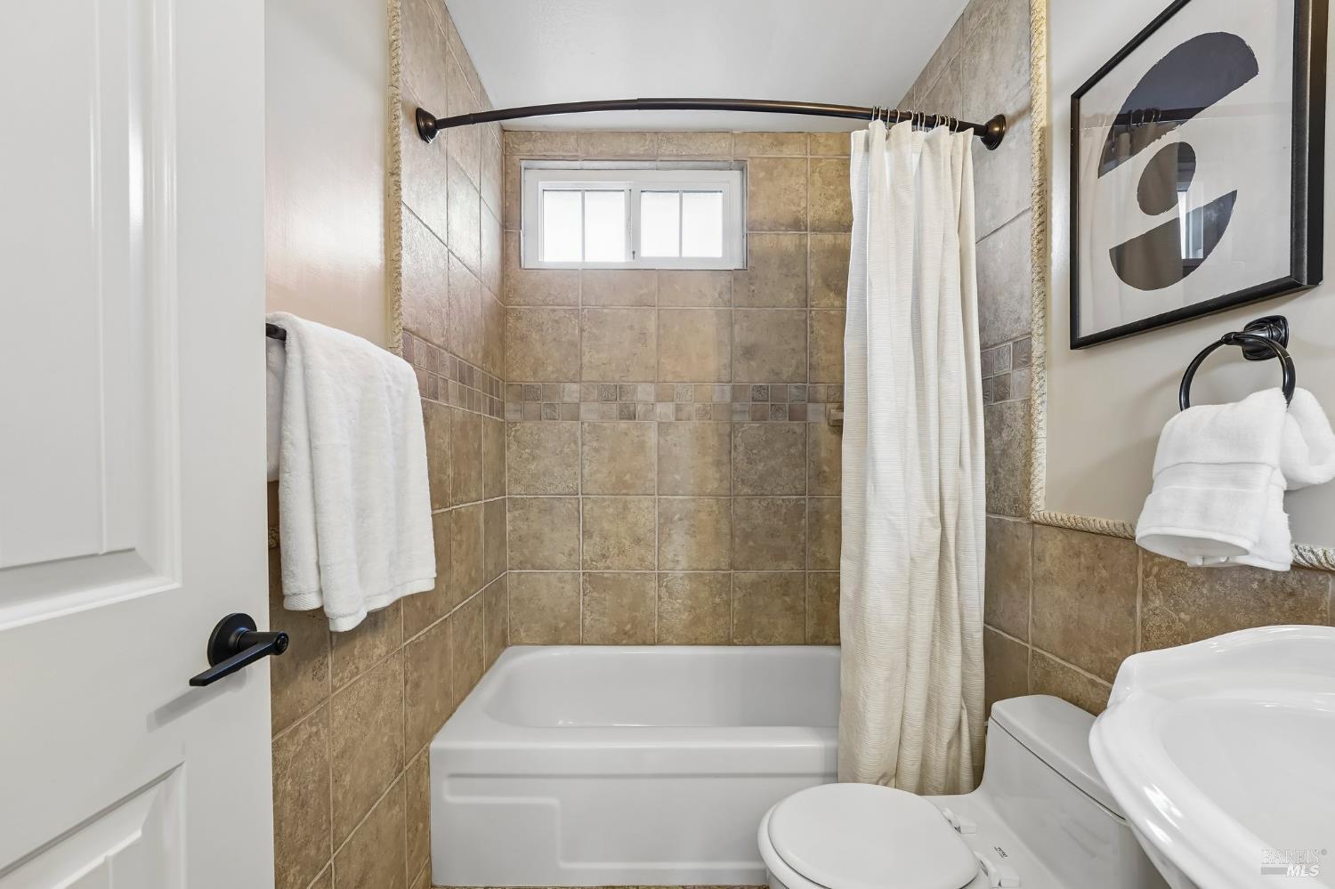 Bathroom with tan tiled tub and shower, white curtain, and a small high window.