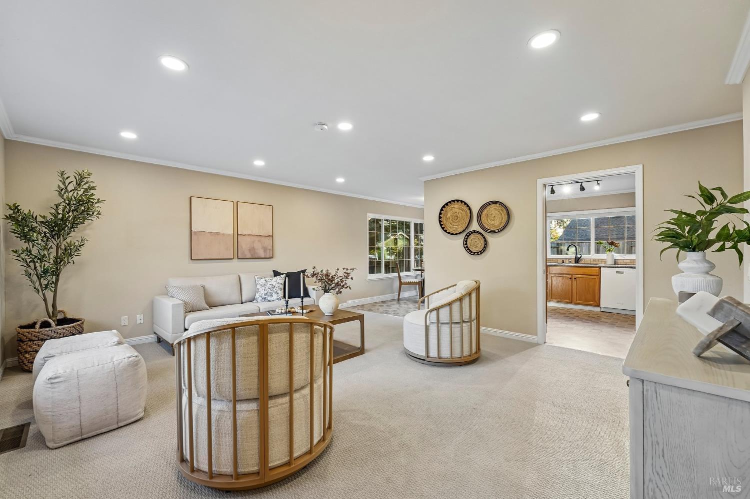 Staged living room with curved wood-framed chairs, cream carpeting, and a view into the kitchen.