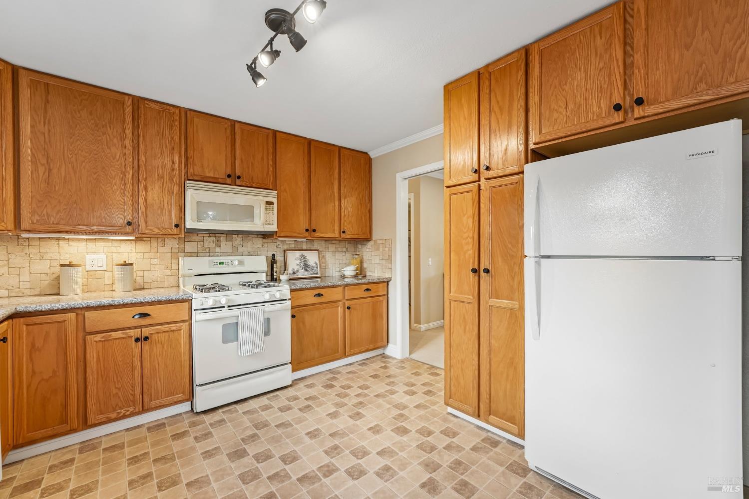 Kitchen with honey-colored wood cabinets, tiled backsplash, white appliances, and patterned floor.