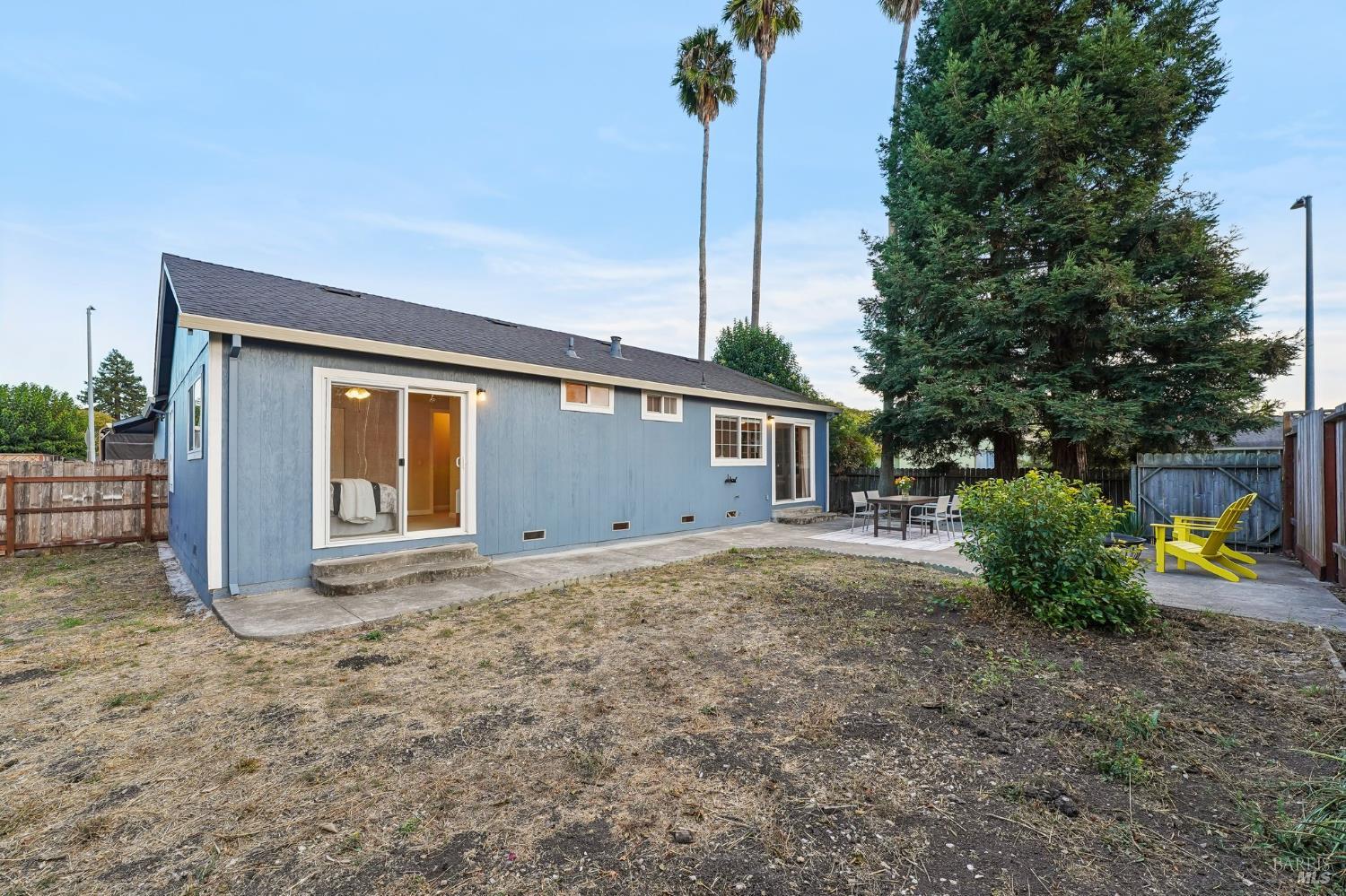 Blue house rear exterior with a patio, yellow Adirondack chairs, and a fenced yard.