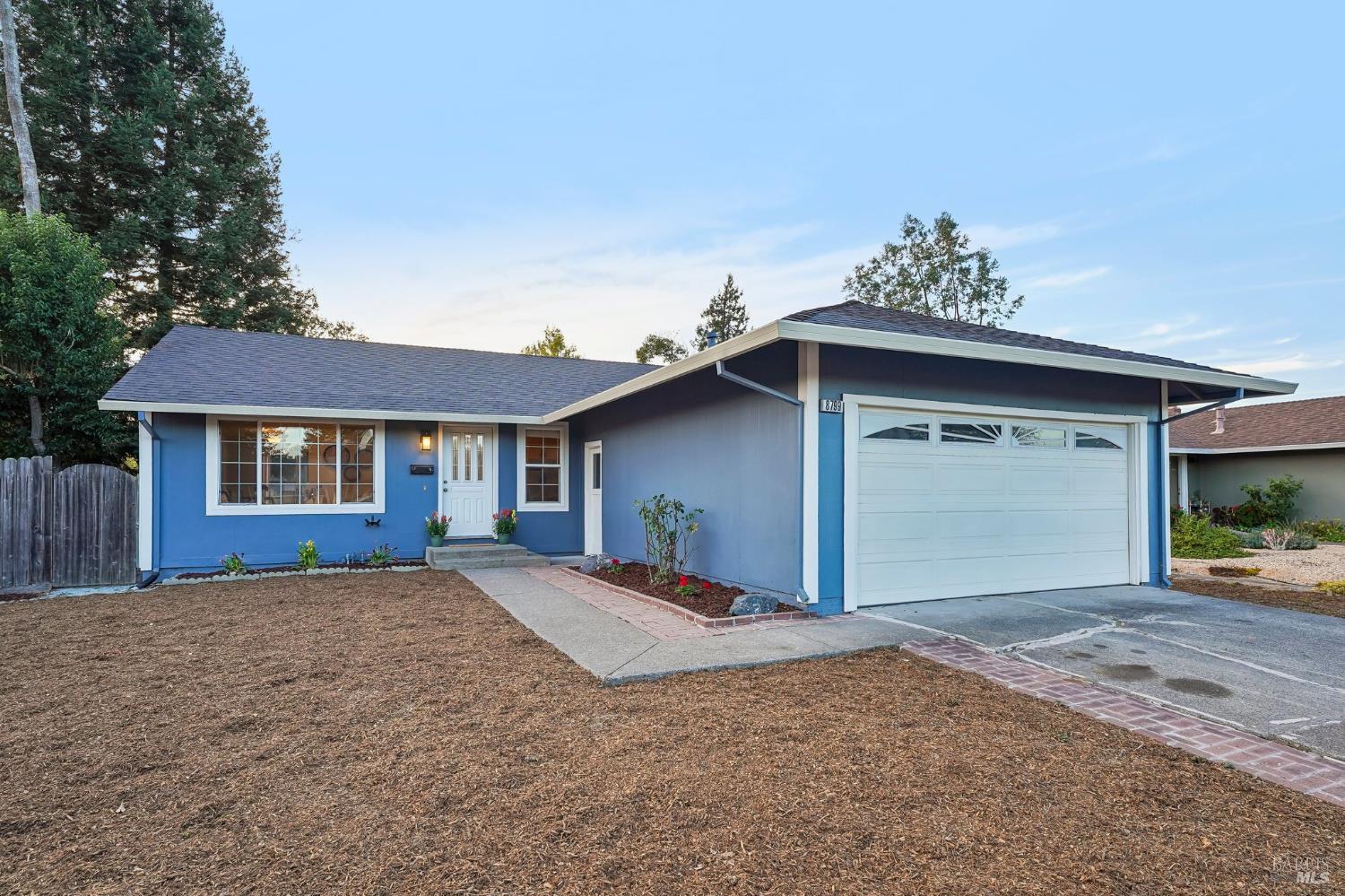 Blue single-story home with a white two-car garage and broad mulch-covered front yard.