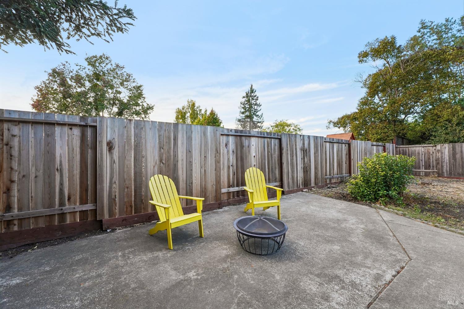 Backyard patio with yellow Adirondack chairs, a round fire pit, and a tall wooden fence.