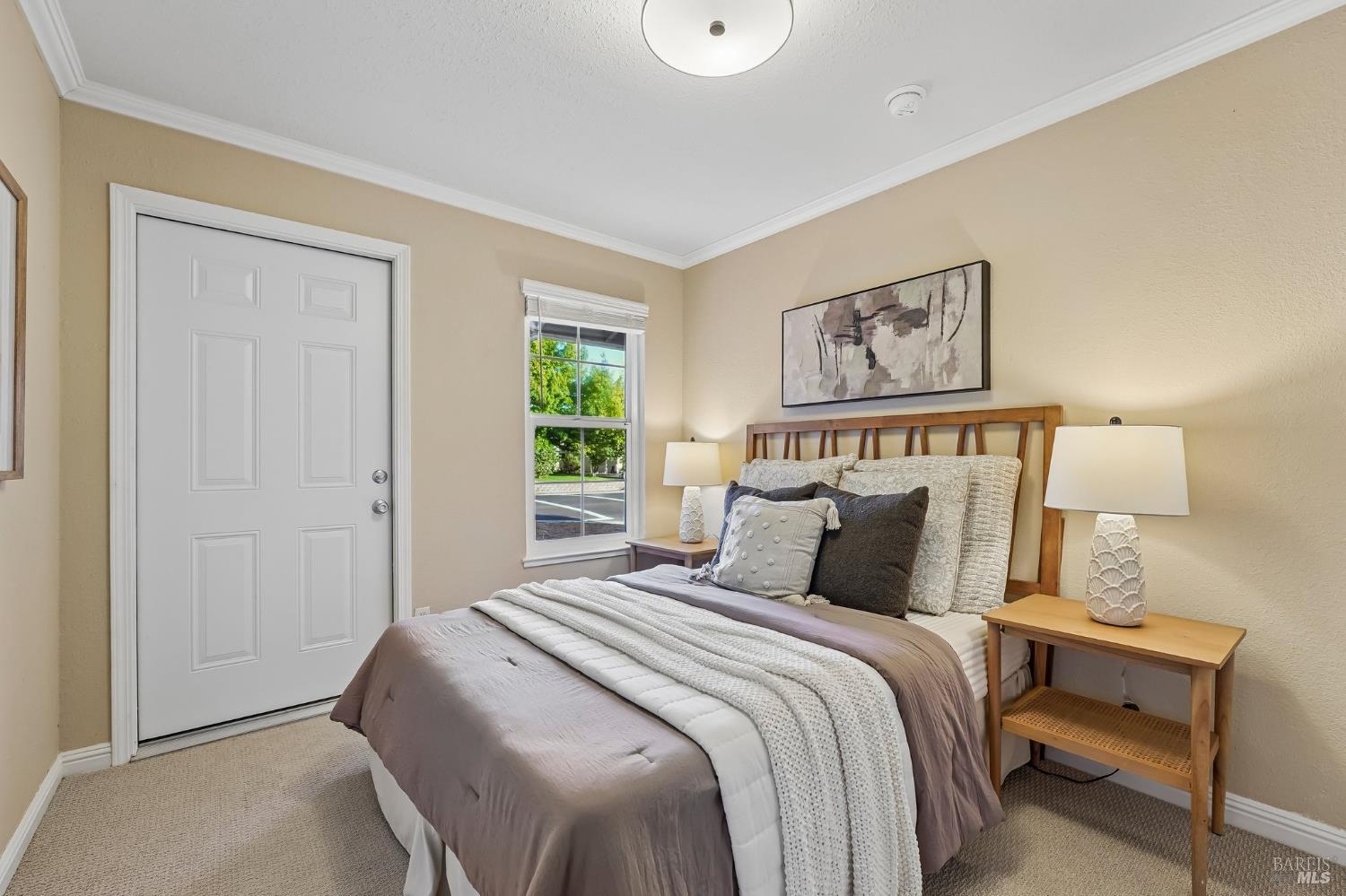 Staged bedroom with layered gray and white bedding, wooden bed frame, and a window overlooking greenery.