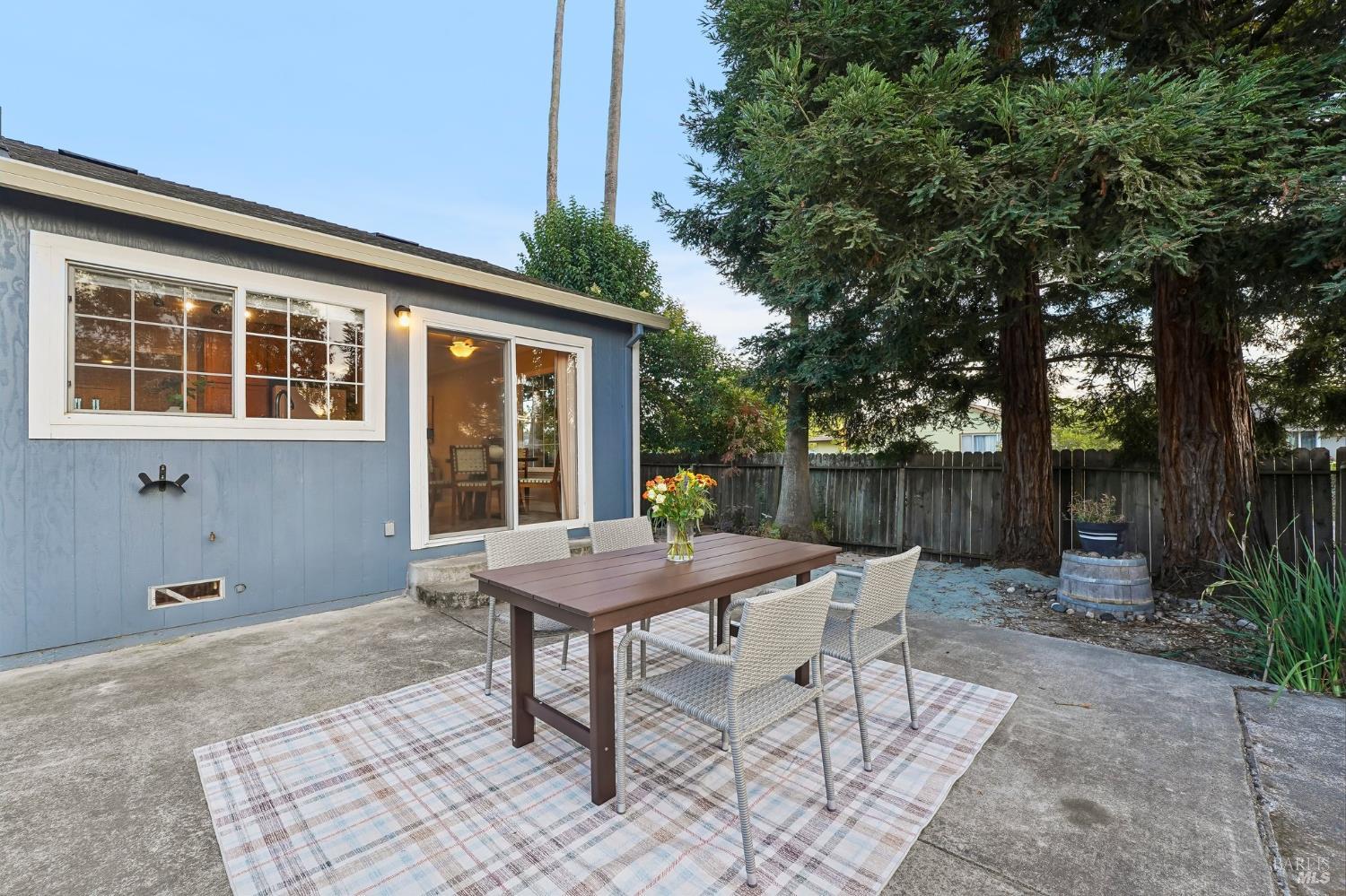 Backyard patio with an outdoor dining set beneath mature trees beside a blue-sided home.