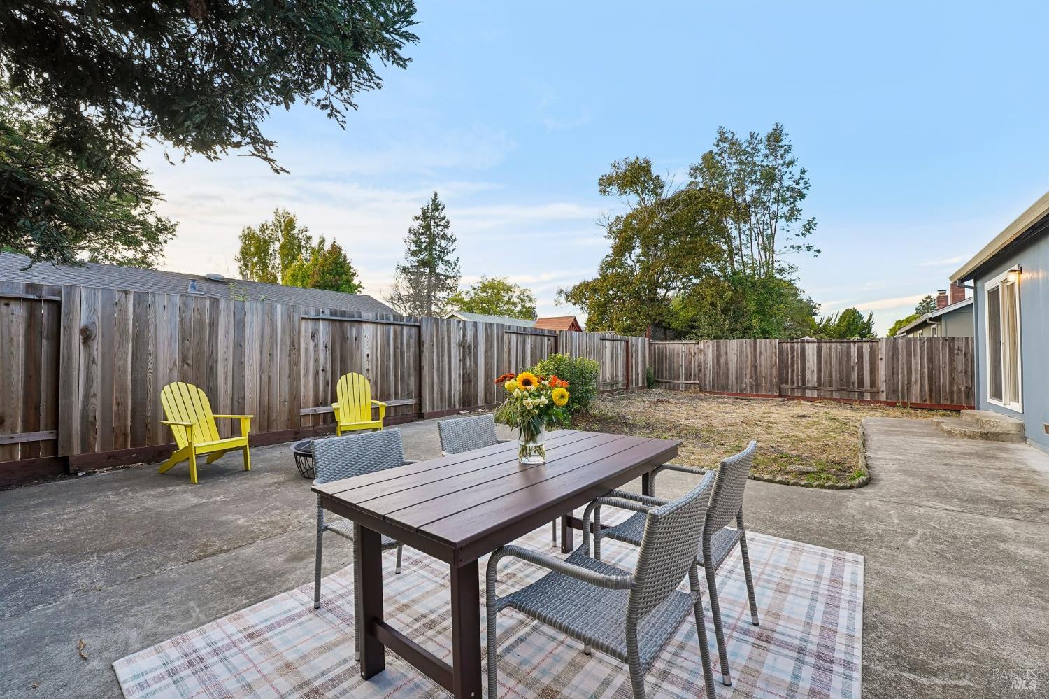Fenced backyard patio with an outdoor dining set, yellow Adirondack chairs, and a dry lawn area.