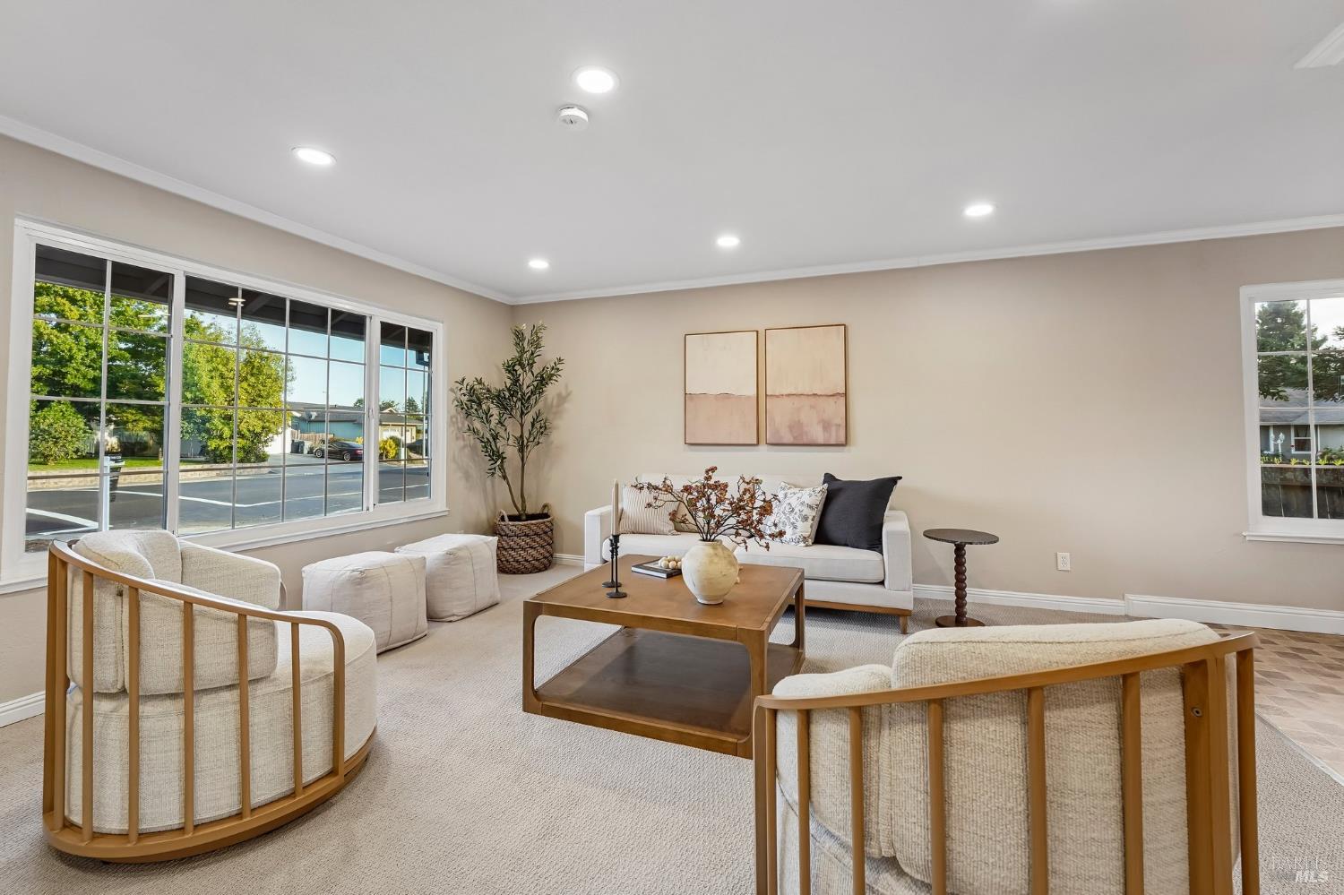 Staged living room with a white sofa, curved wood-frame chairs, and large front windows.