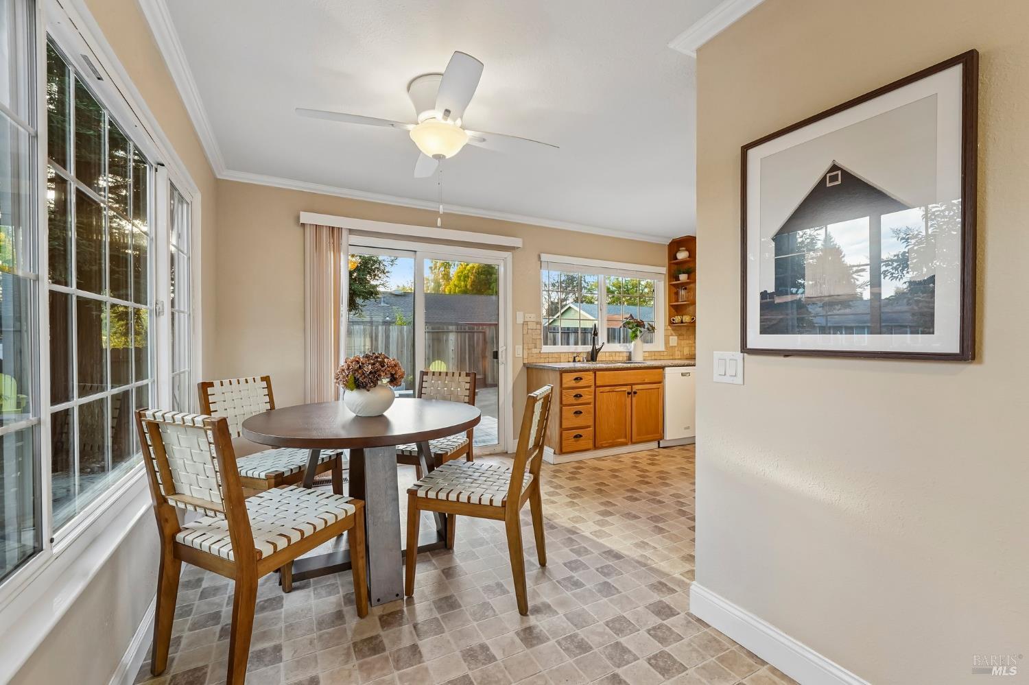 Dining area with a round table and woven chairs beside large windows and an open kitchen.