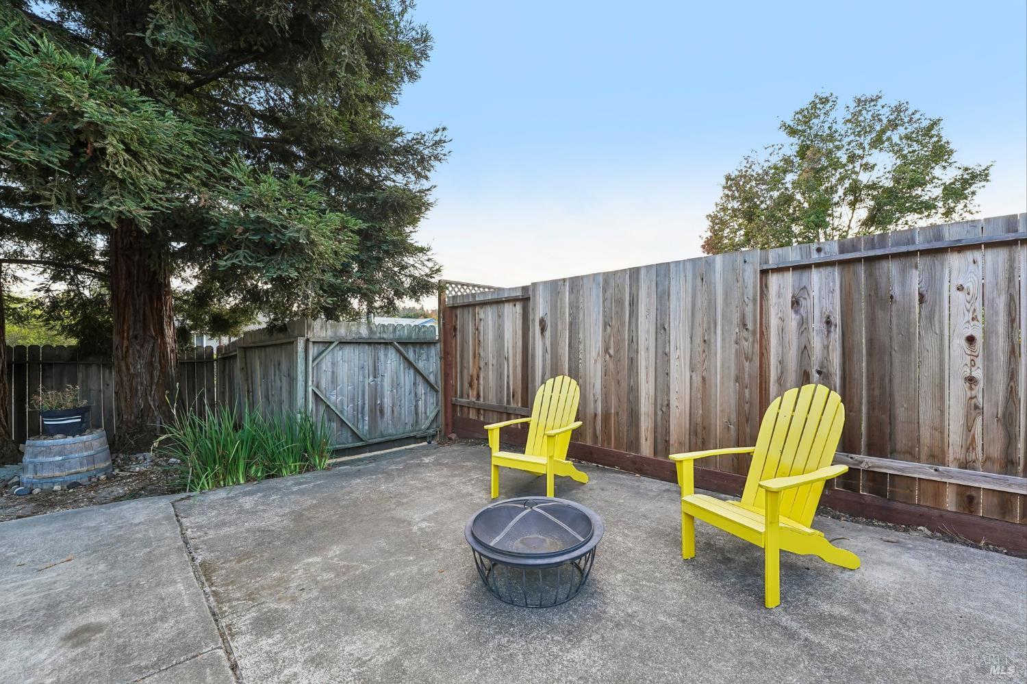 Fenced concrete backyard patio with yellow Adirondack chairs, a metal fire pit, and a large evergreen tree.