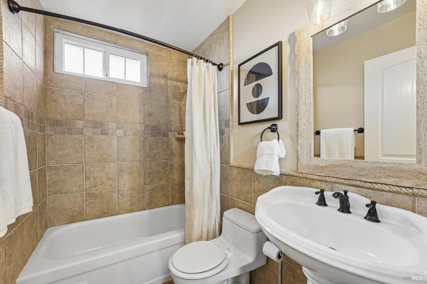 Bathroom with tan tiled tub and shower, pedestal sink, large mirror, and high window.