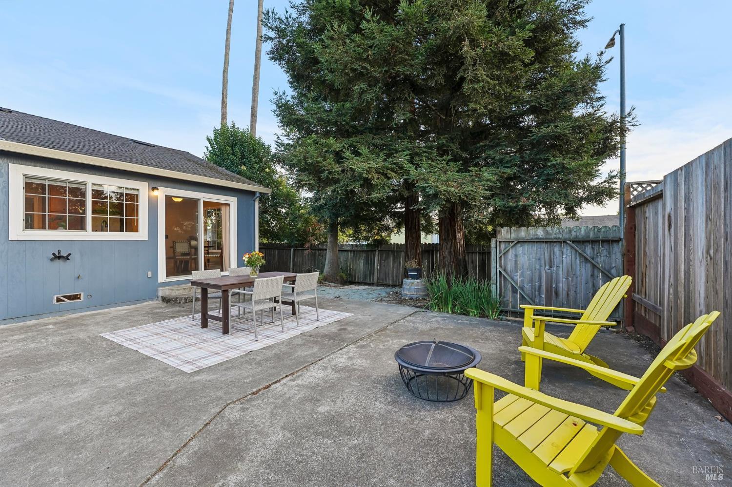 Fenced backyard patio with an outdoor dining set, yellow Adirondack chairs, and mature trees.