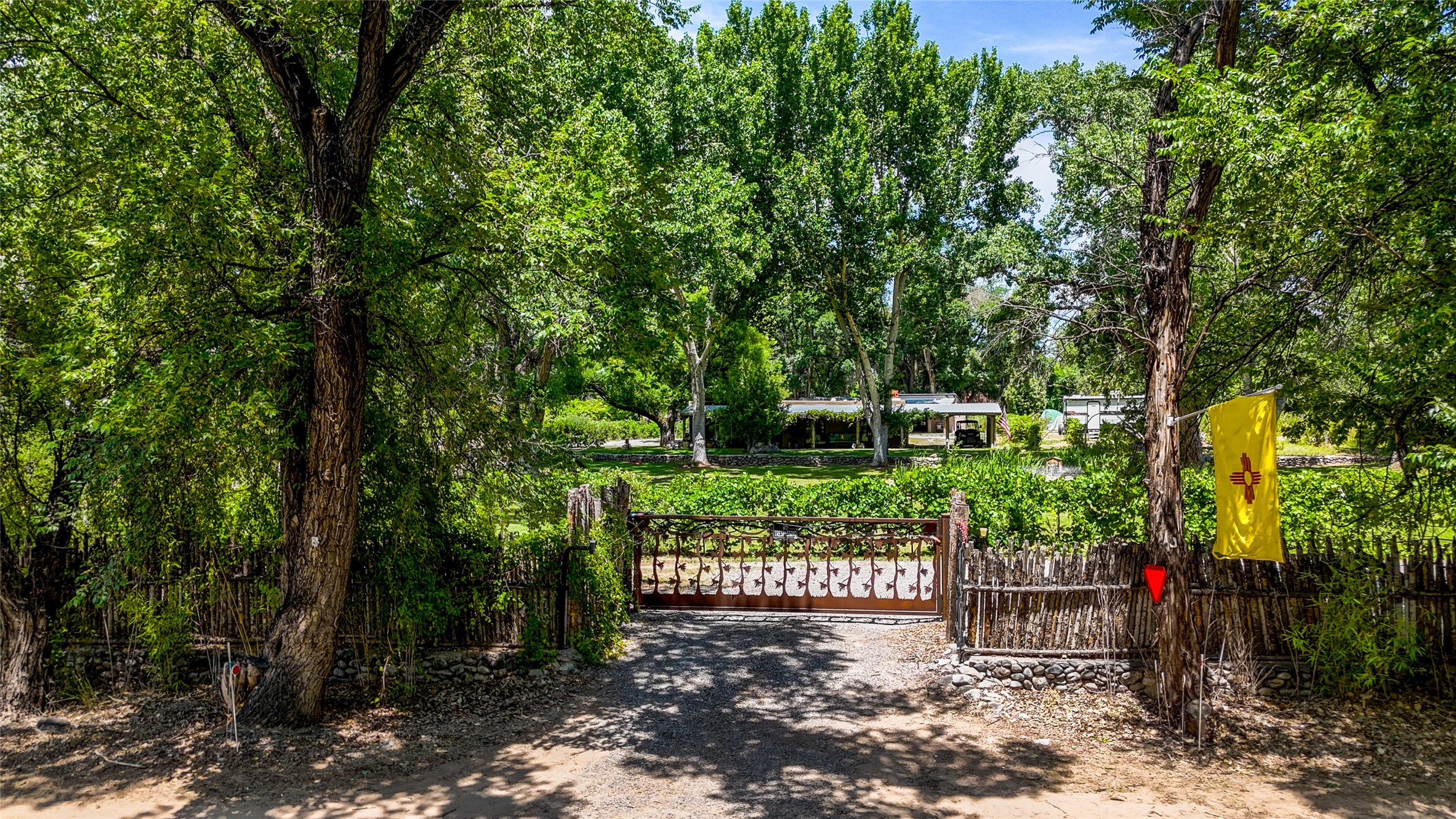 Tree-lined gravel driveway and decorative gate leading to a home, with a New Mexico flag beside the fence.