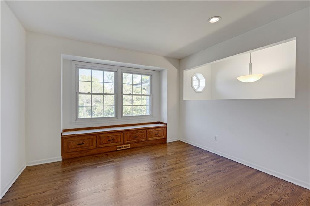 Empty room with hardwood floors, a built-in wooden window seat, and a wide window overlooking trees.