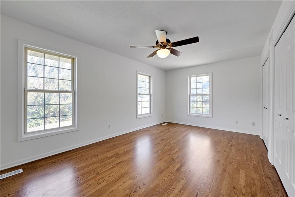 Empty bedroom with hardwood floors, three windows, a ceiling fan, and white closets.