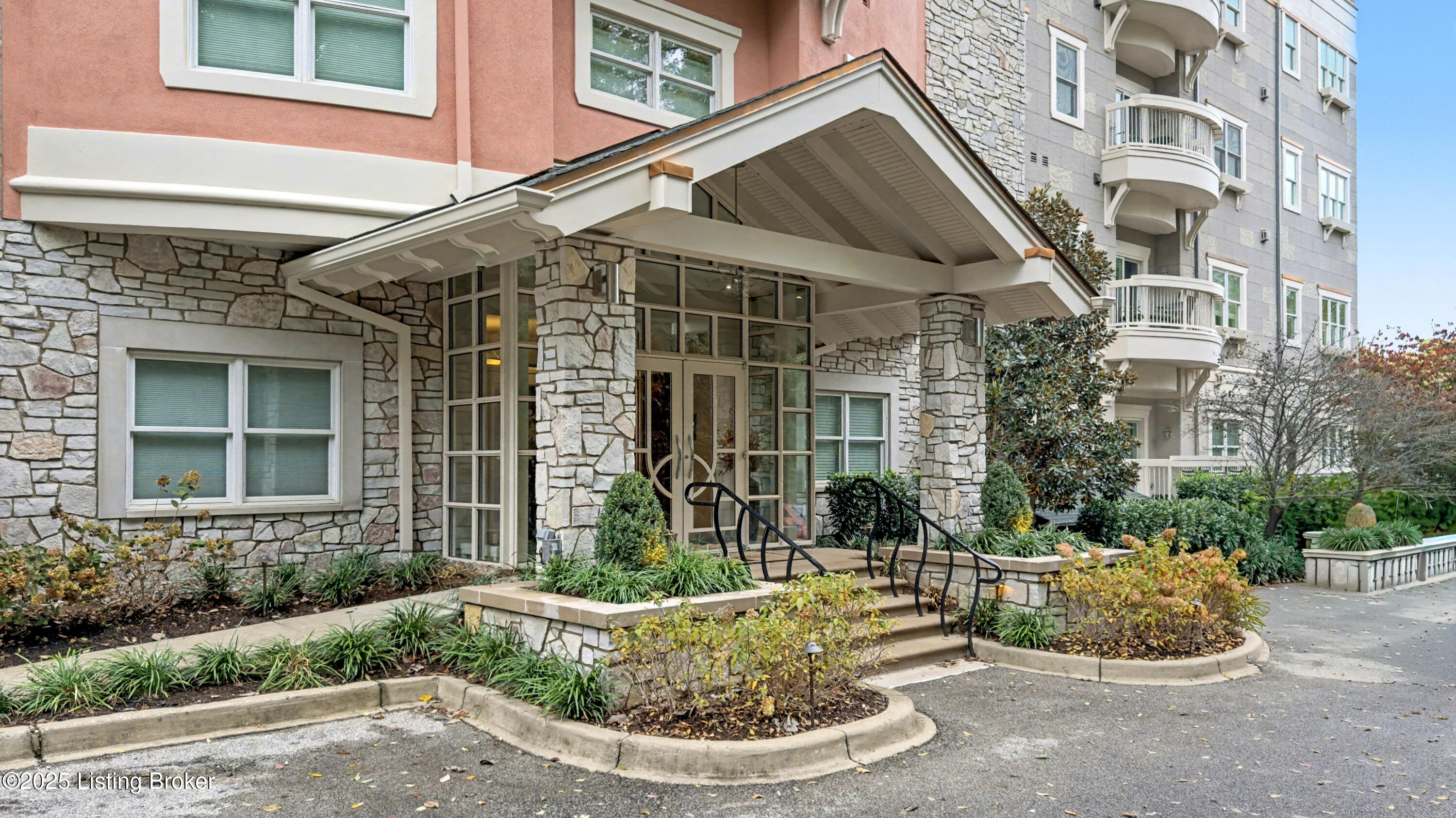 Residential building entrance with a stone facade, covered portico, landscaped beds, and curved balconies.