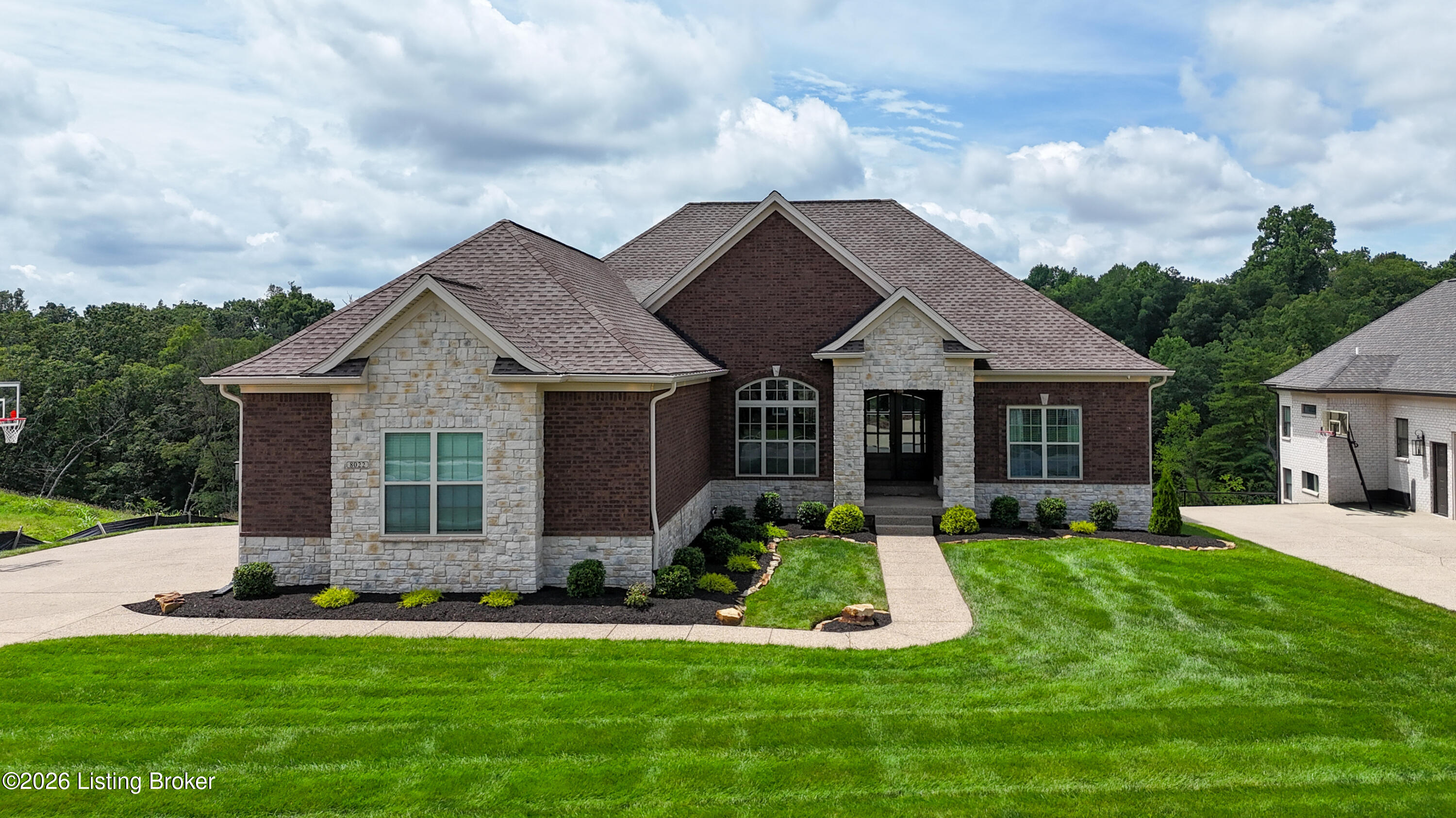 Brick and stone house with a multi-gabled roof, arched window, landscaped beds, and broad lawn.