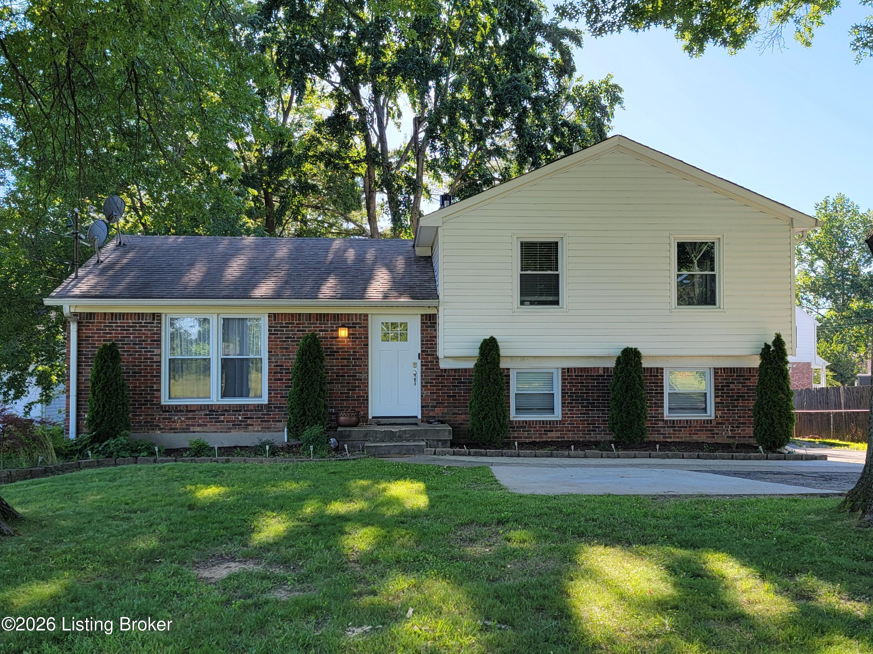 Split-level brick and white-sided home beneath mature trees, with a broad lawn and driveway.
