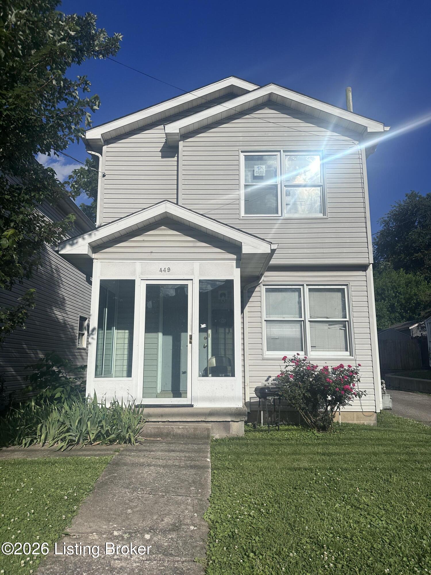 Two-story gray-sided house with a glass entry porch, lawn, and flowering shrubs.