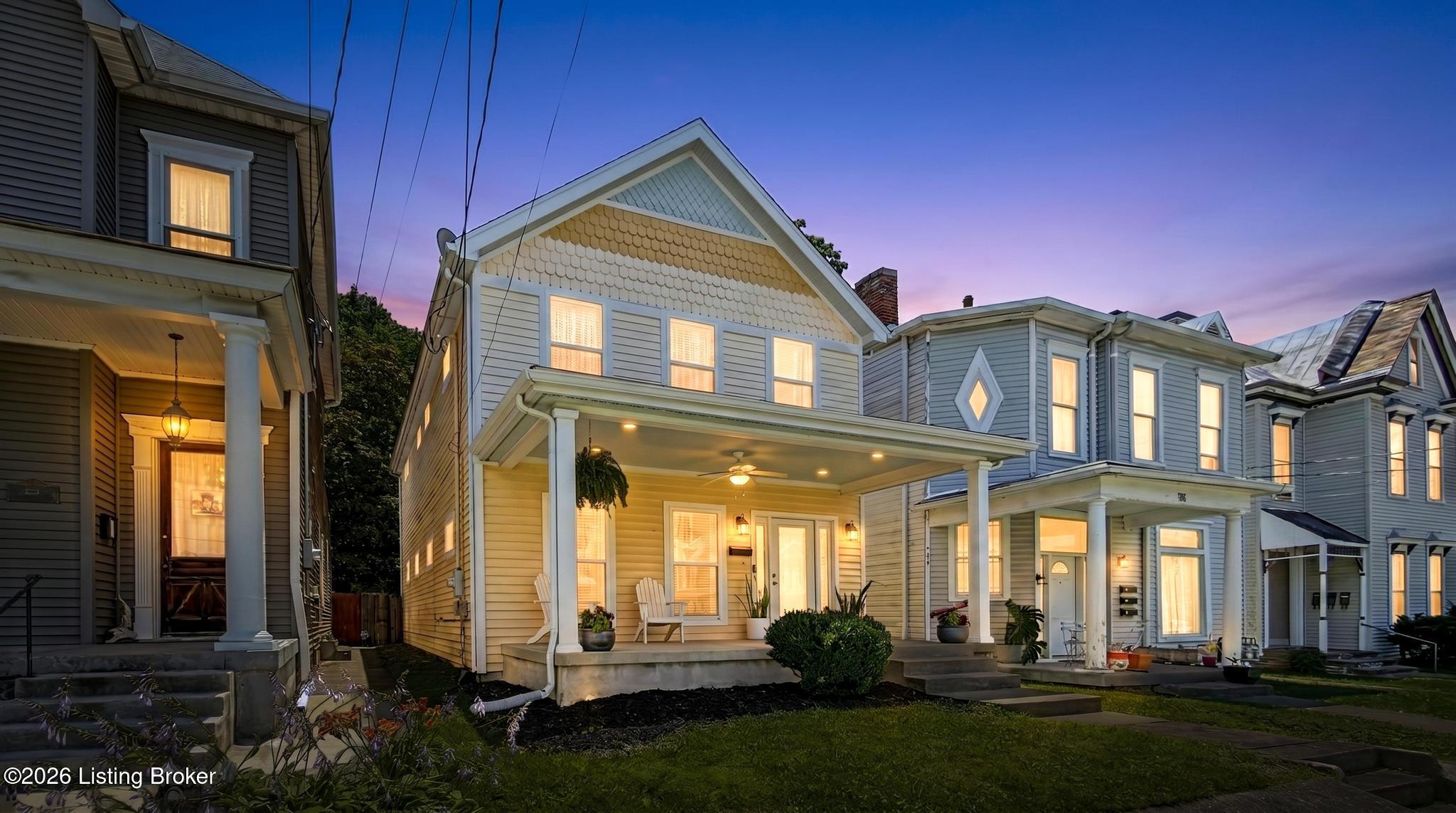 A light-sided house with a broad covered porch glows at dusk among neighboring homes.