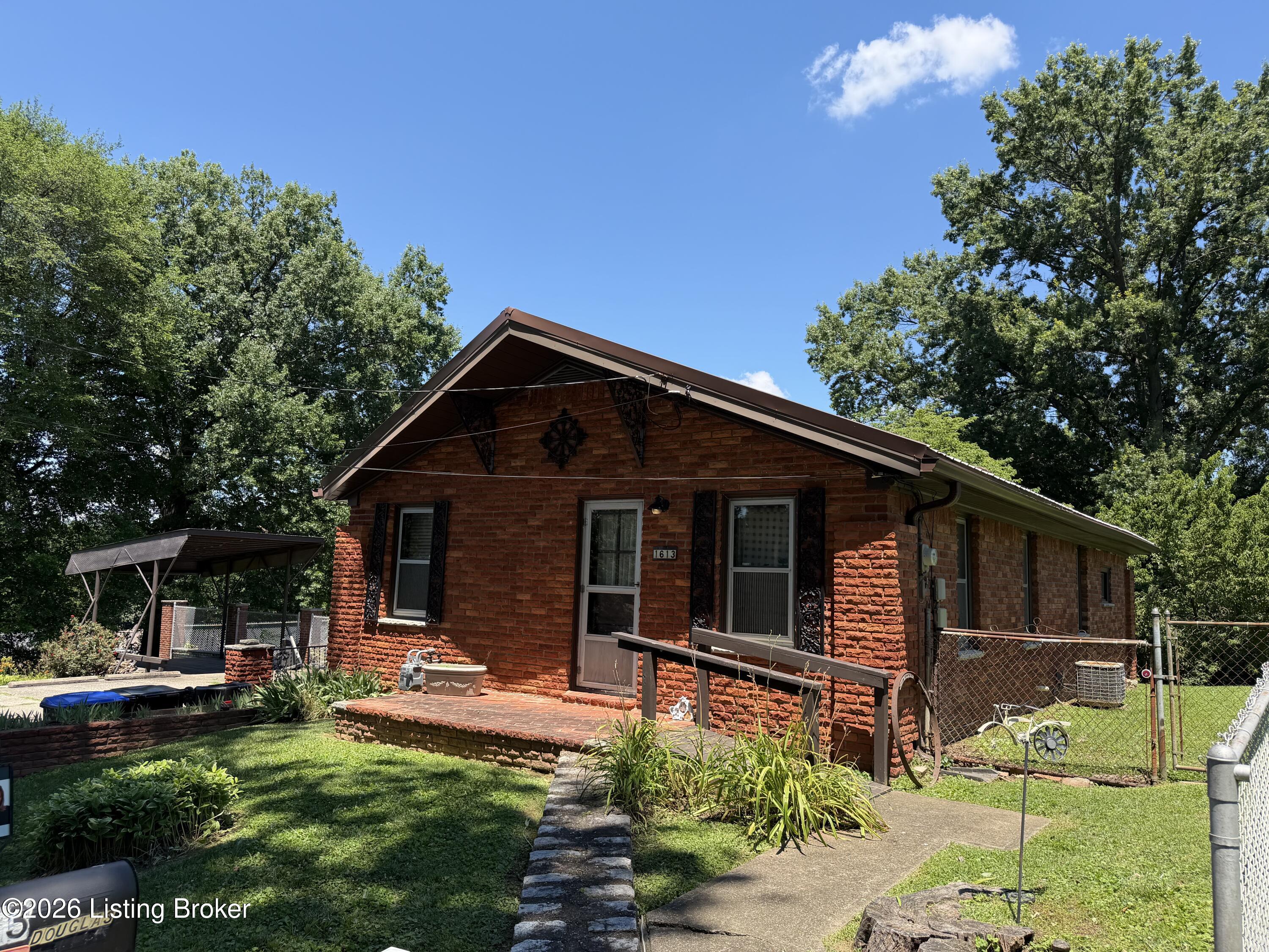 Red brick house with a front porch, fenced yard, and covered carport beneath mature trees.