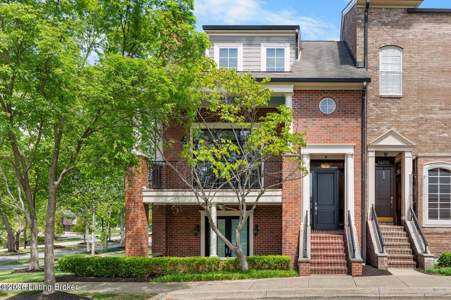 Red-brick townhouse with a covered balcony, front steps, and leafy trees.