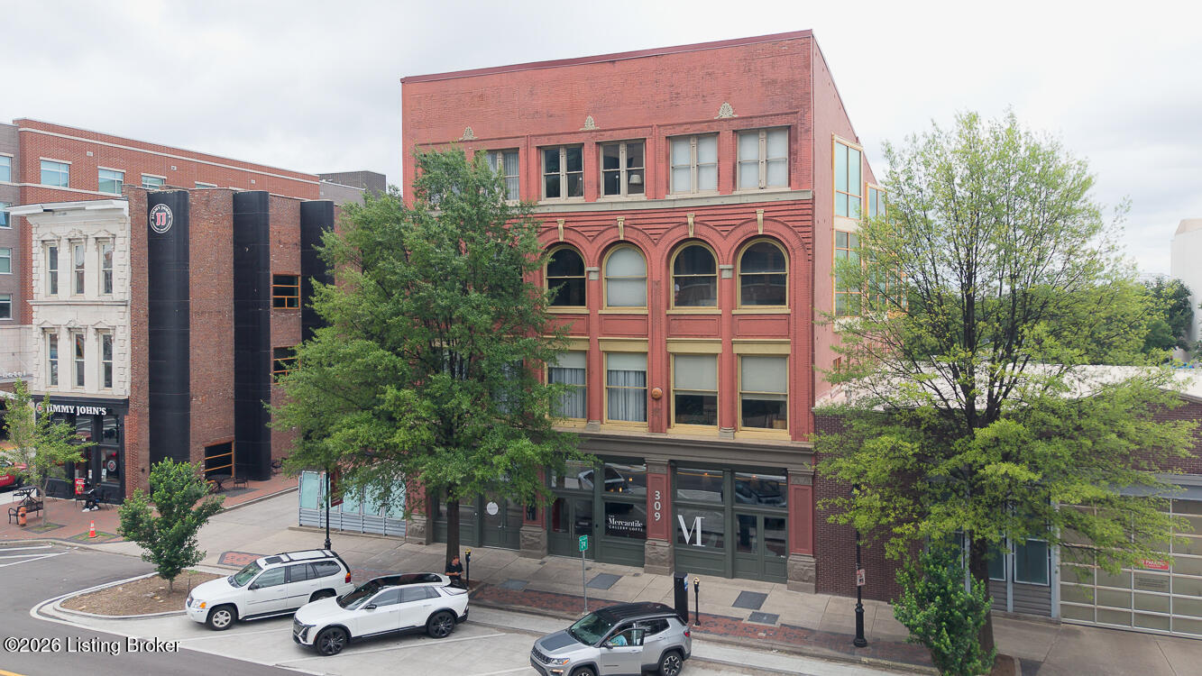 Red-brick commercial building with arched windows and storefronts, framed by leafy trees.