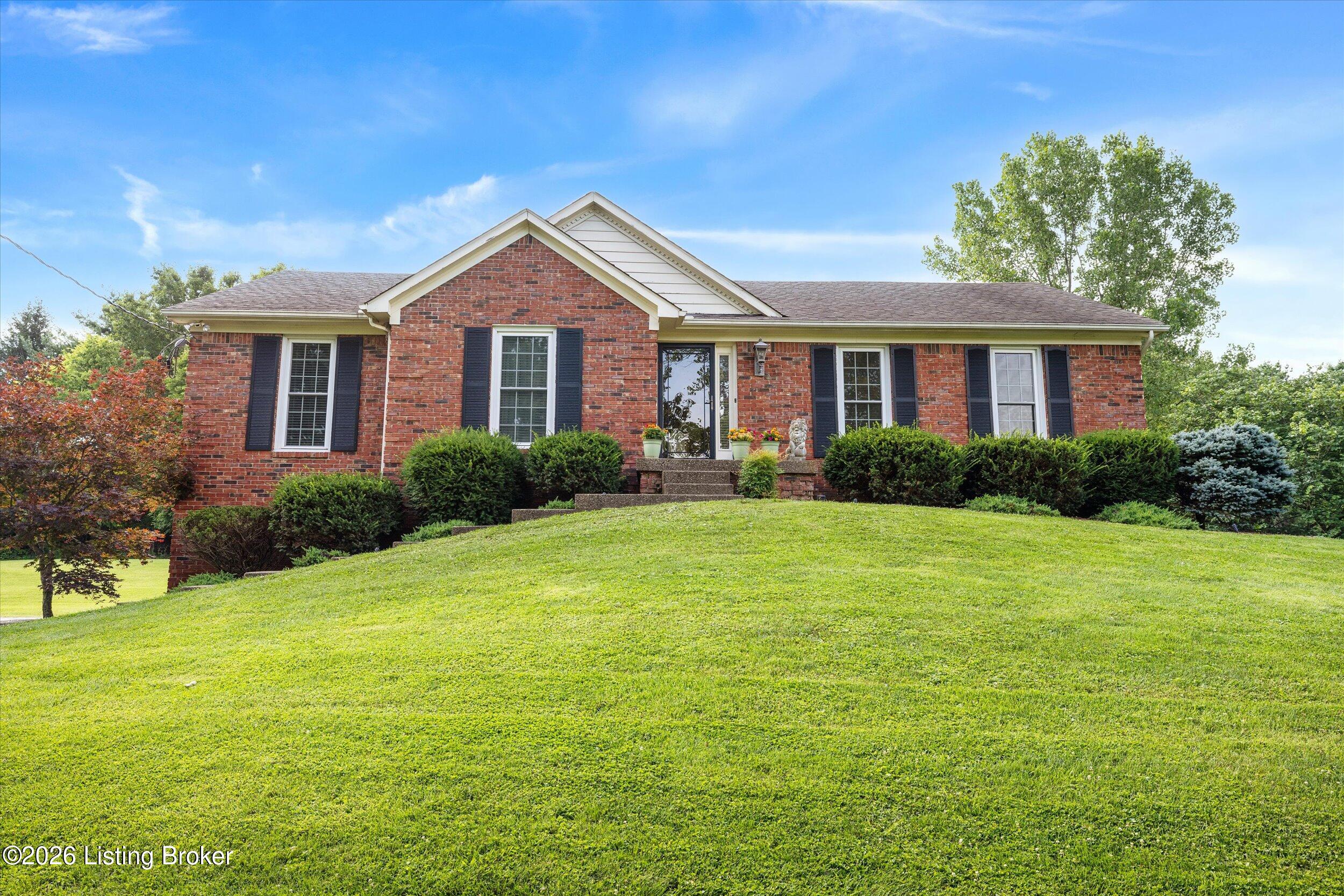 Red brick single-story home with dark shutters, landscaped shrubs, and a broad sloping lawn.