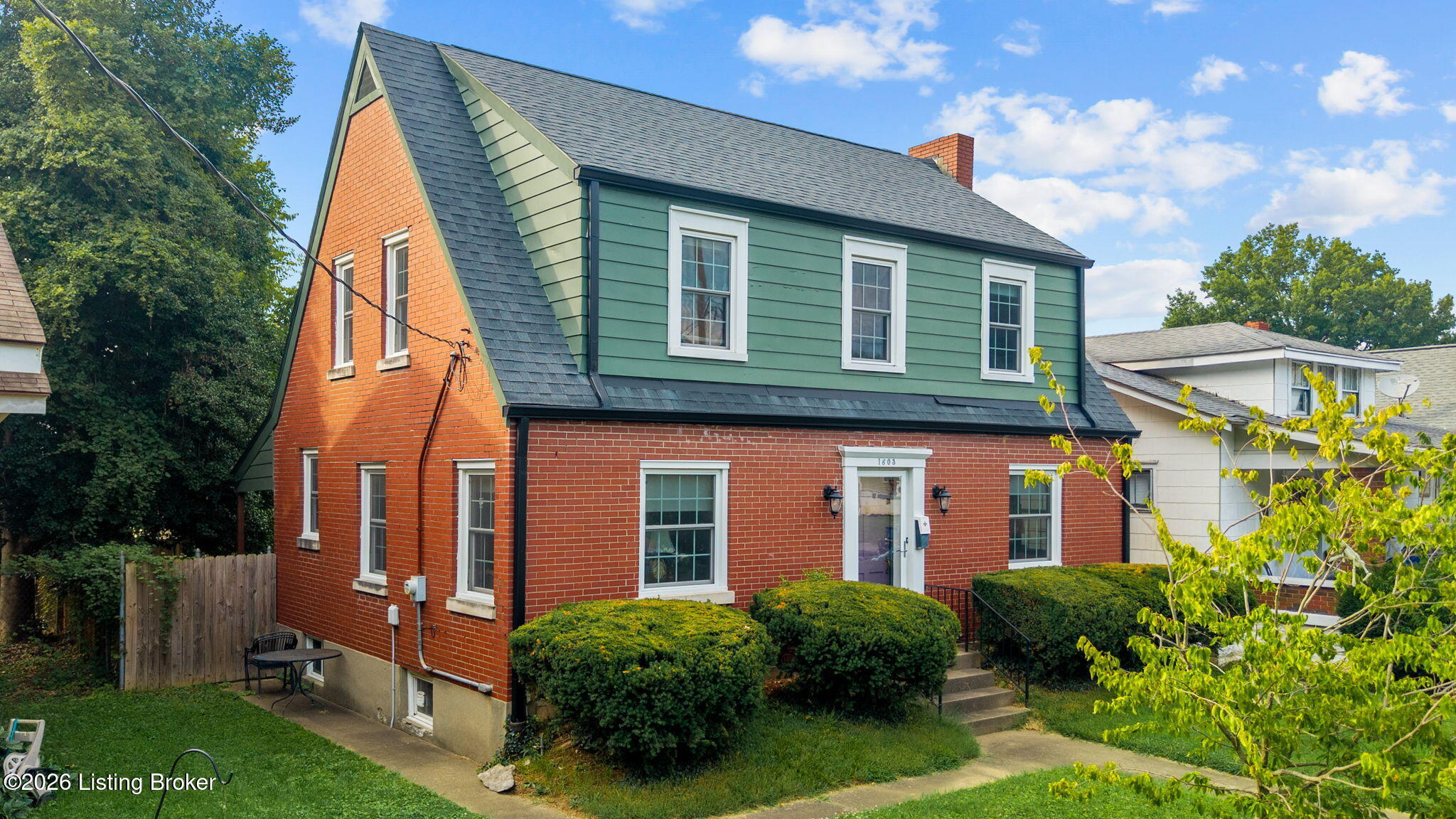 Two-story brick house with green siding, steep gabled roof, and trimmed front shrubs.