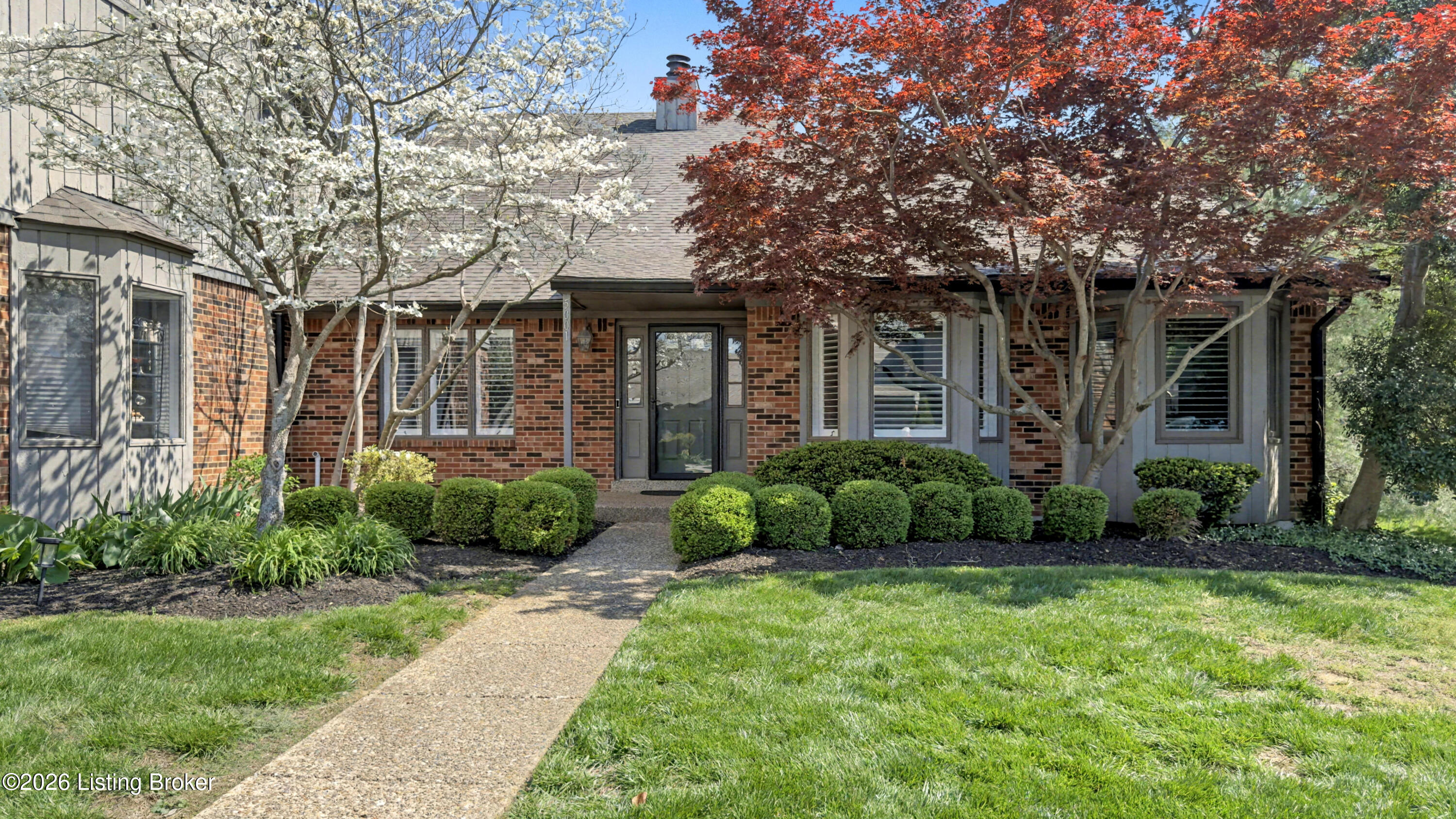 Brick home framed by flowering and red-leaved trees, with a curved walkway and manicured shrubs.