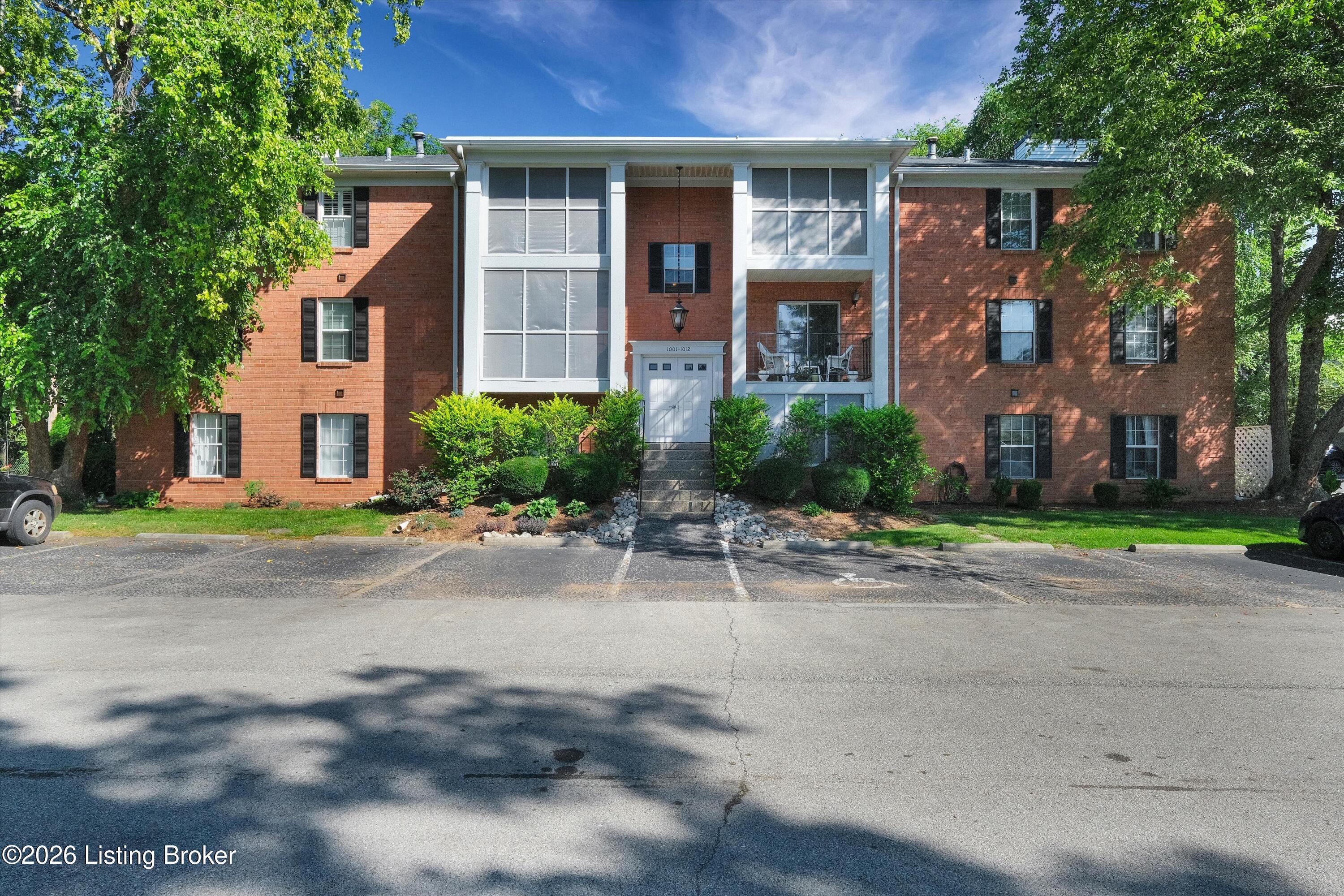 Red-brick apartment building with a central entrance, white trim, and mature trees along the front.