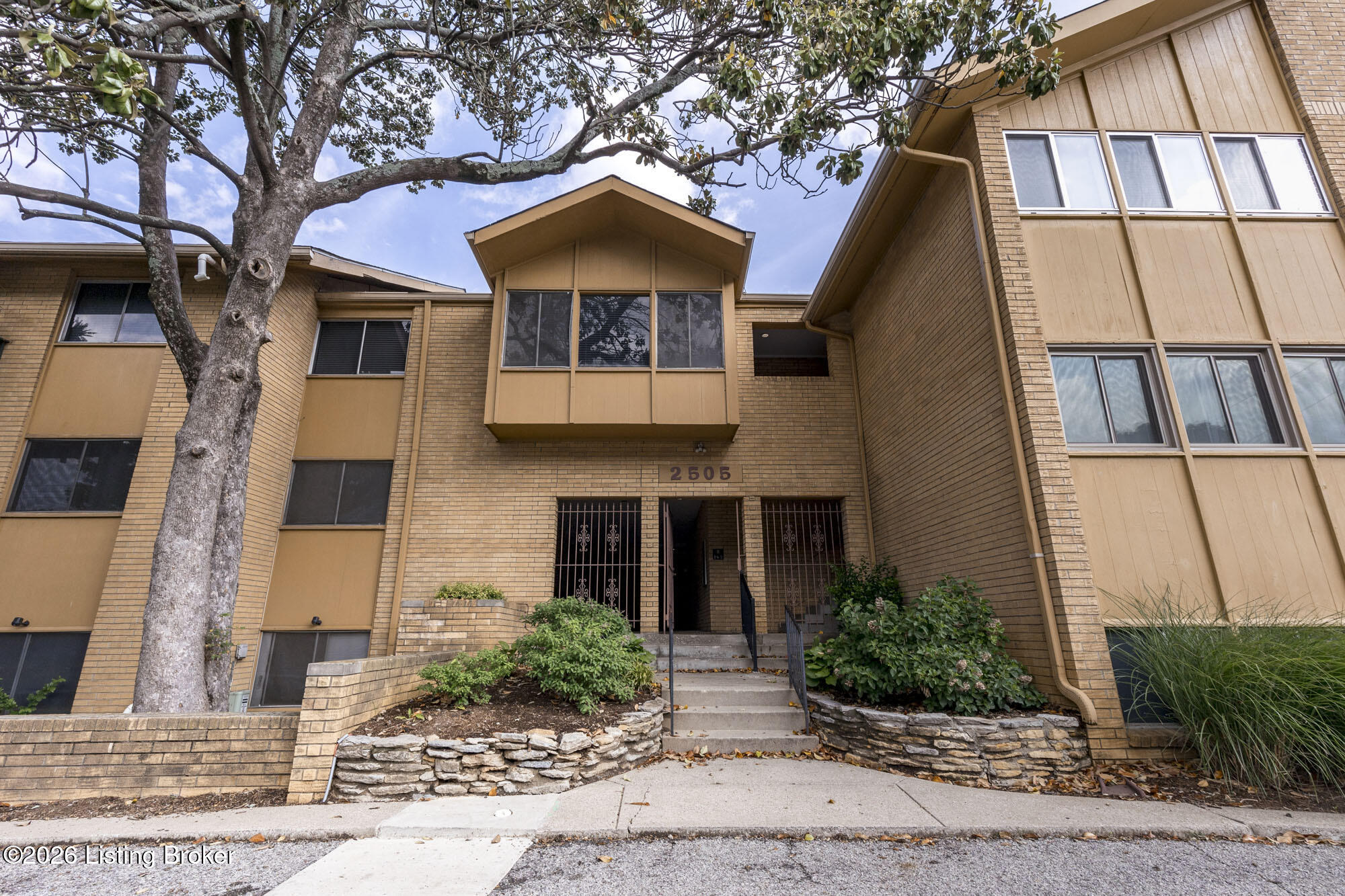 Brick apartment building entrance with a central projecting bay, stone planters, and a large tree.