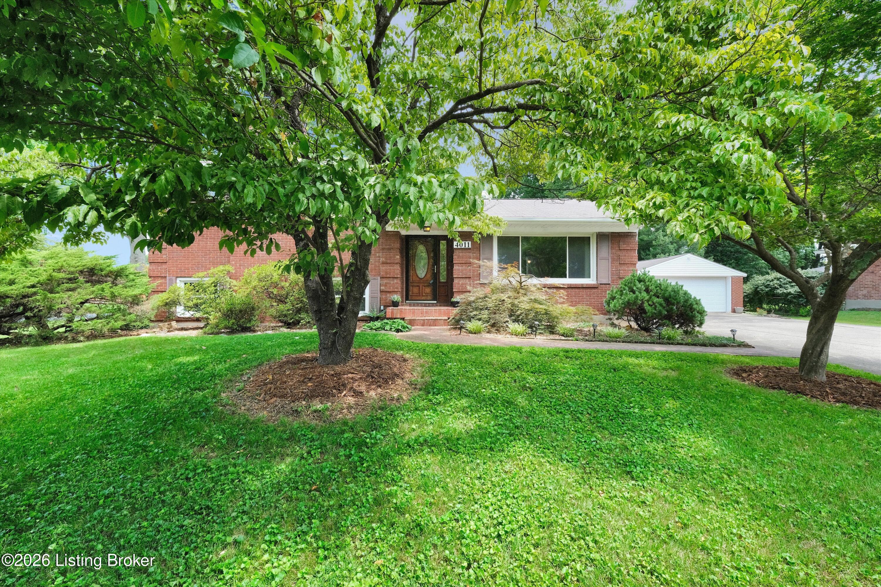 Red-brick house with a white garage, mature trees, and a broad green lawn.