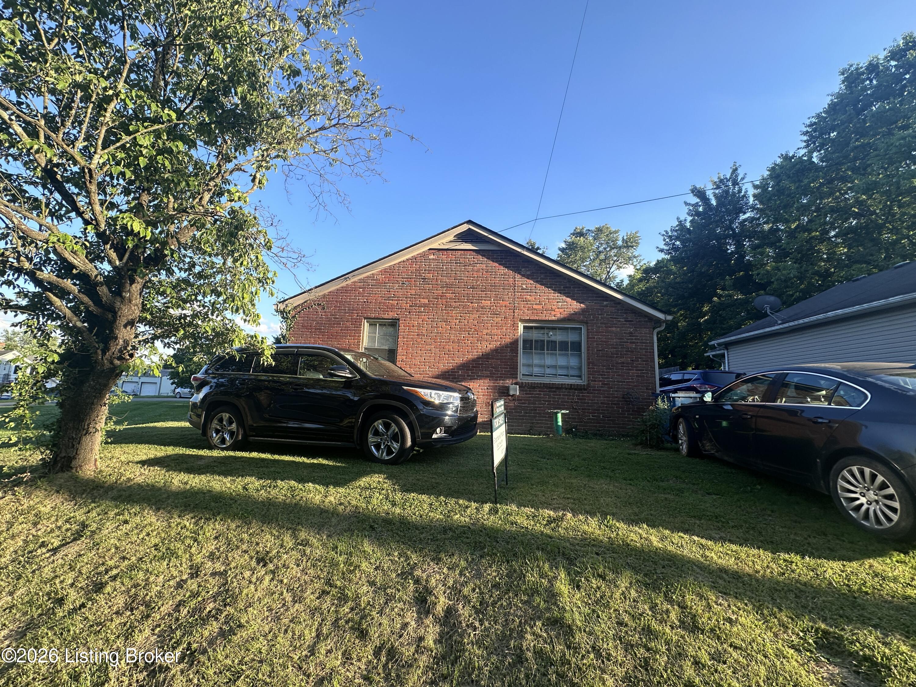 Red brick house with a front lawn, mature trees, and two parked cars.