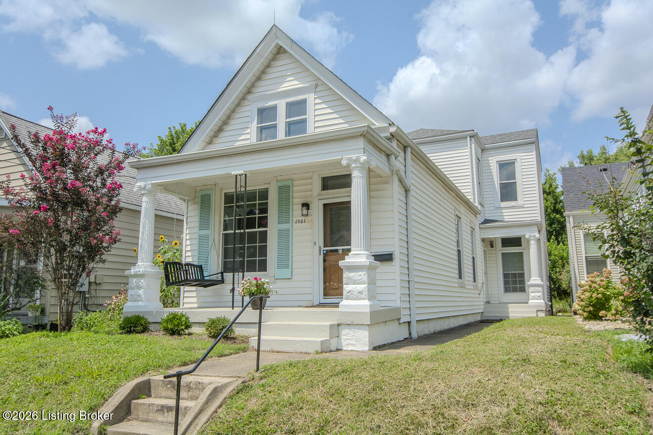White gabled house with a columned porch, pale green shutters, and flowering shrubs.