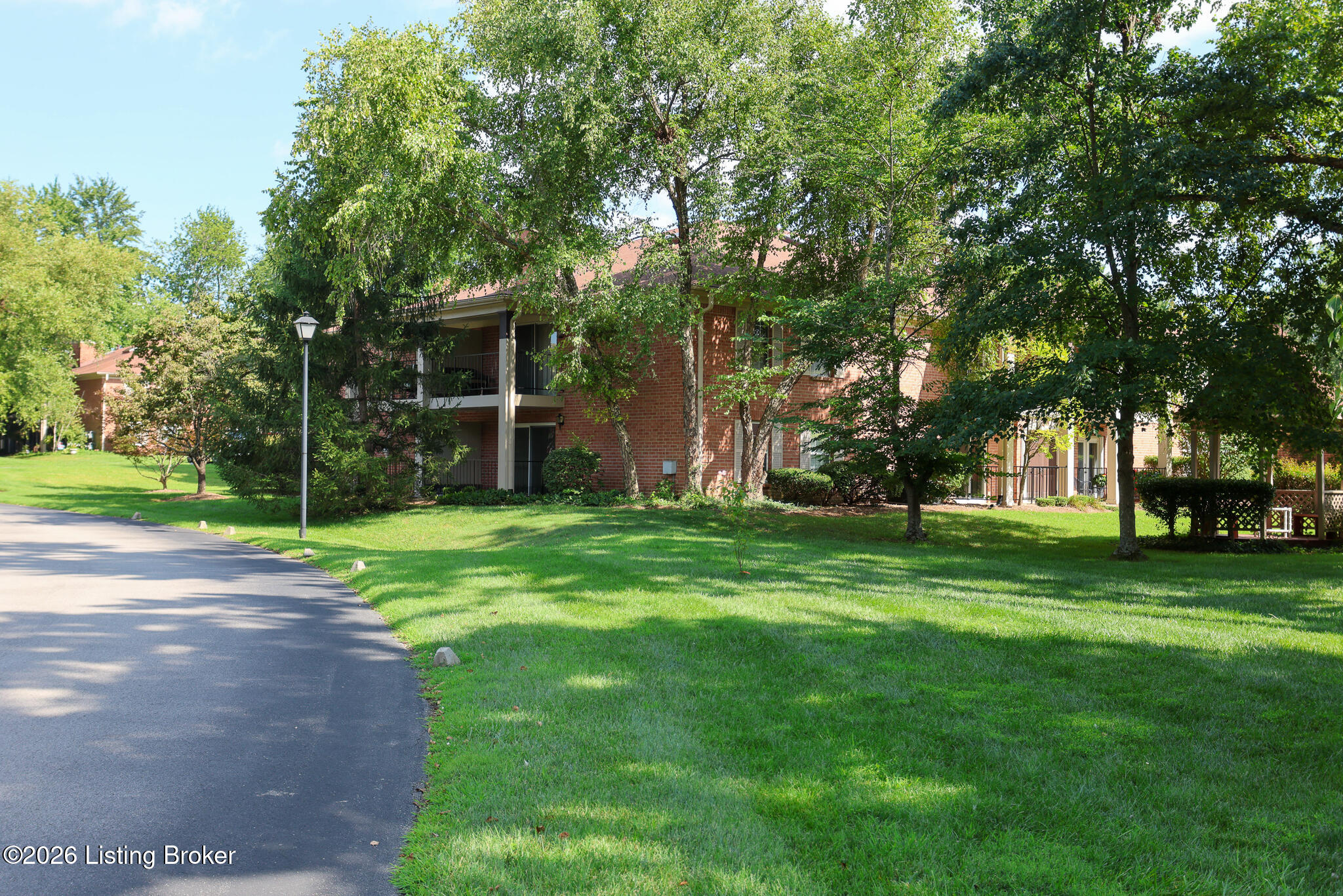Red-brick apartment building shaded by mature trees, with balconies, a green lawn, and a curving driveway.